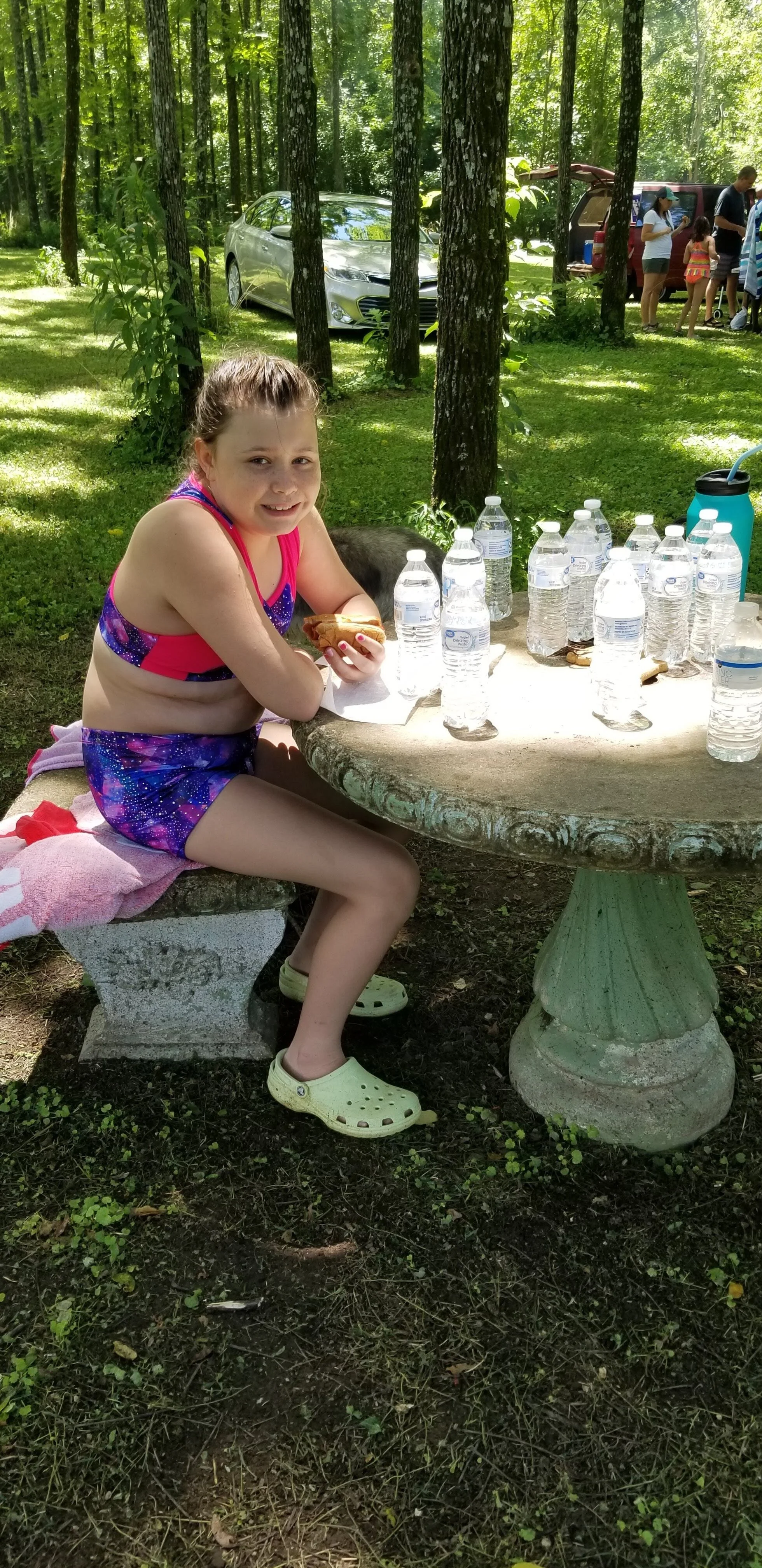 Child in colorful swimsuit sitting at stone table with water bottles, holding a sandwich, surrounded by a forest setting.