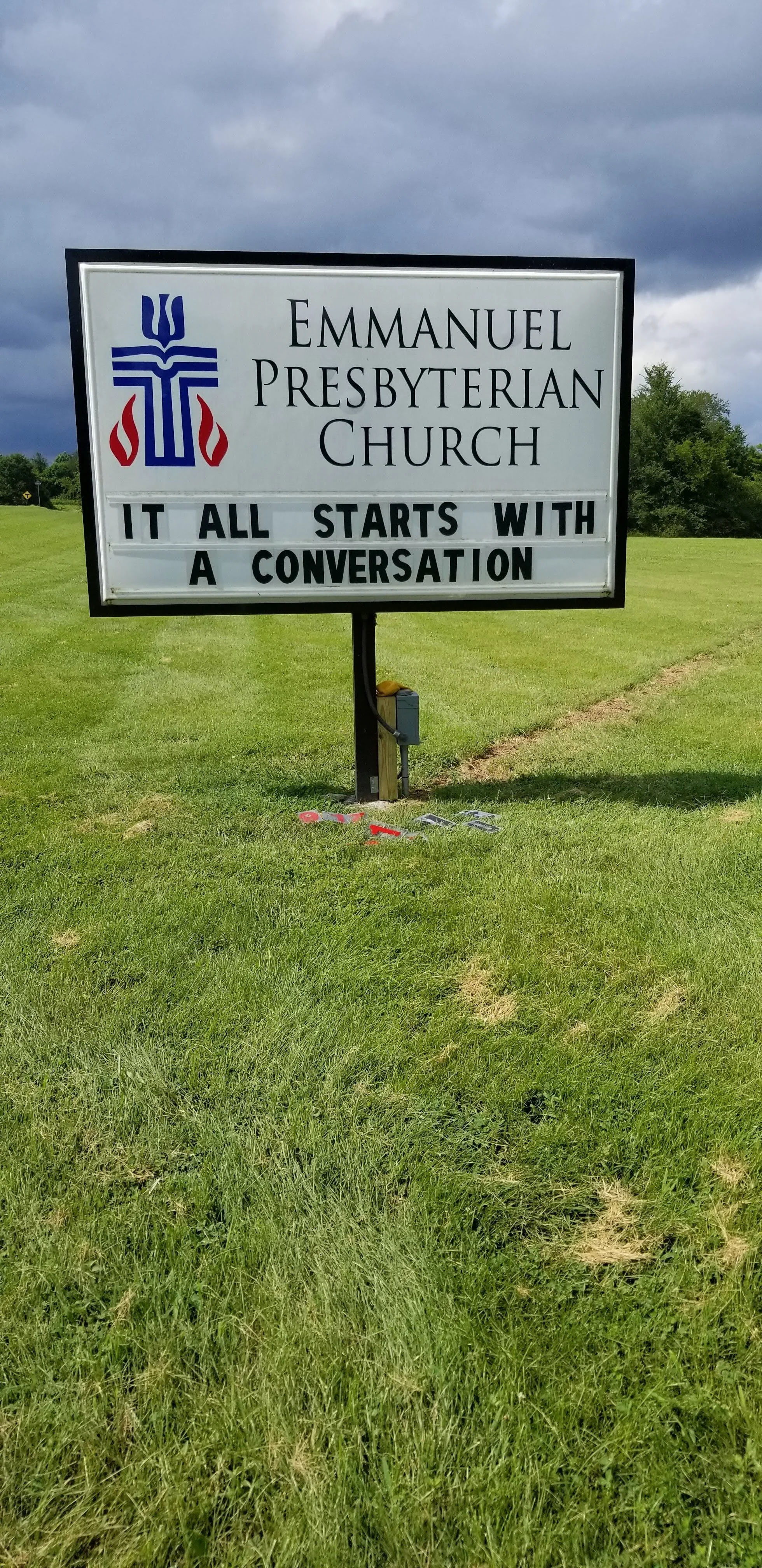 Church sign reading "Emmanuel Presbyterian Church. It all starts with a conversation" on a grassy lawn with cloudy sky.
