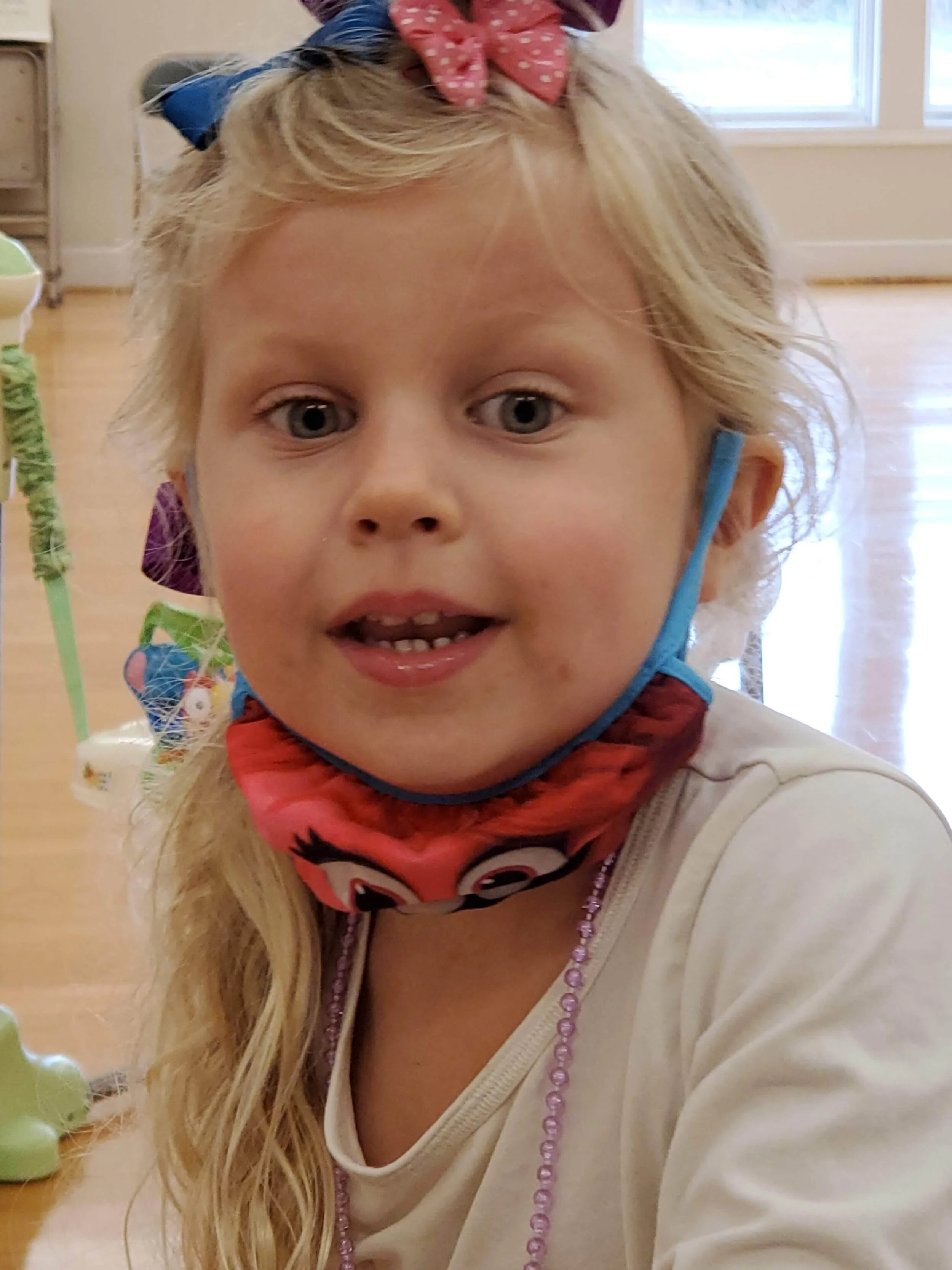 Young girl with long blonde hair, wearing a colorful bow and a face mask under her chin.
