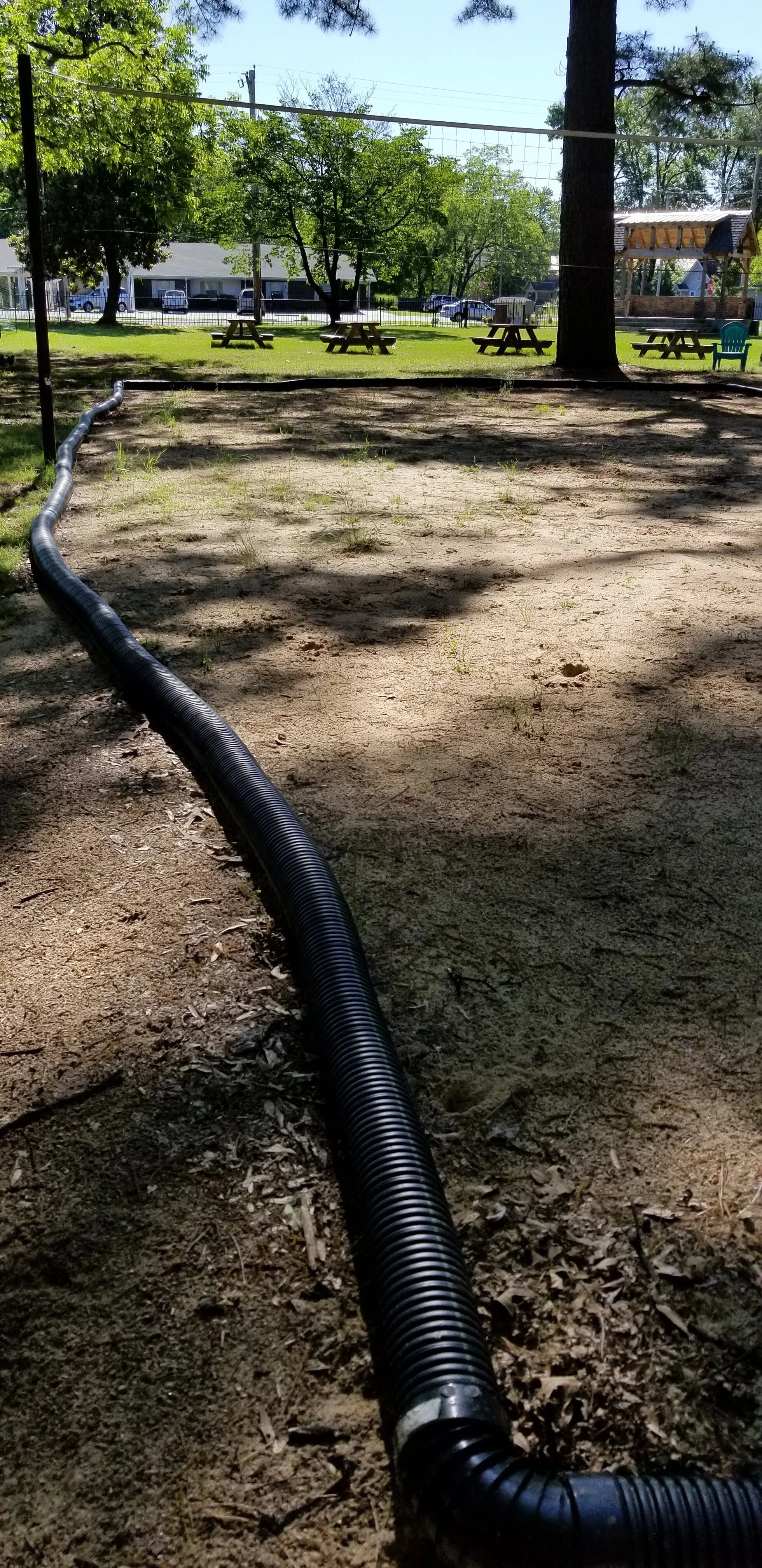 Outdoor volleyball court with a black drainage pipe on the sand surface, surrounded by trees and picnic tables in a park setting.