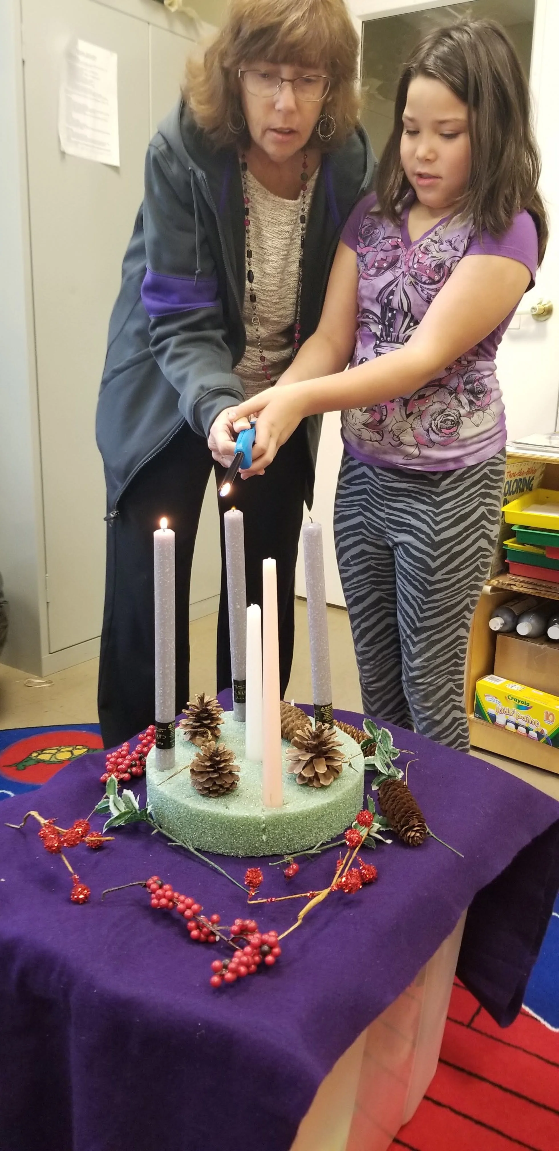A woman and a girl lighting Advent candles on a decorated stand with pine cones and berries.