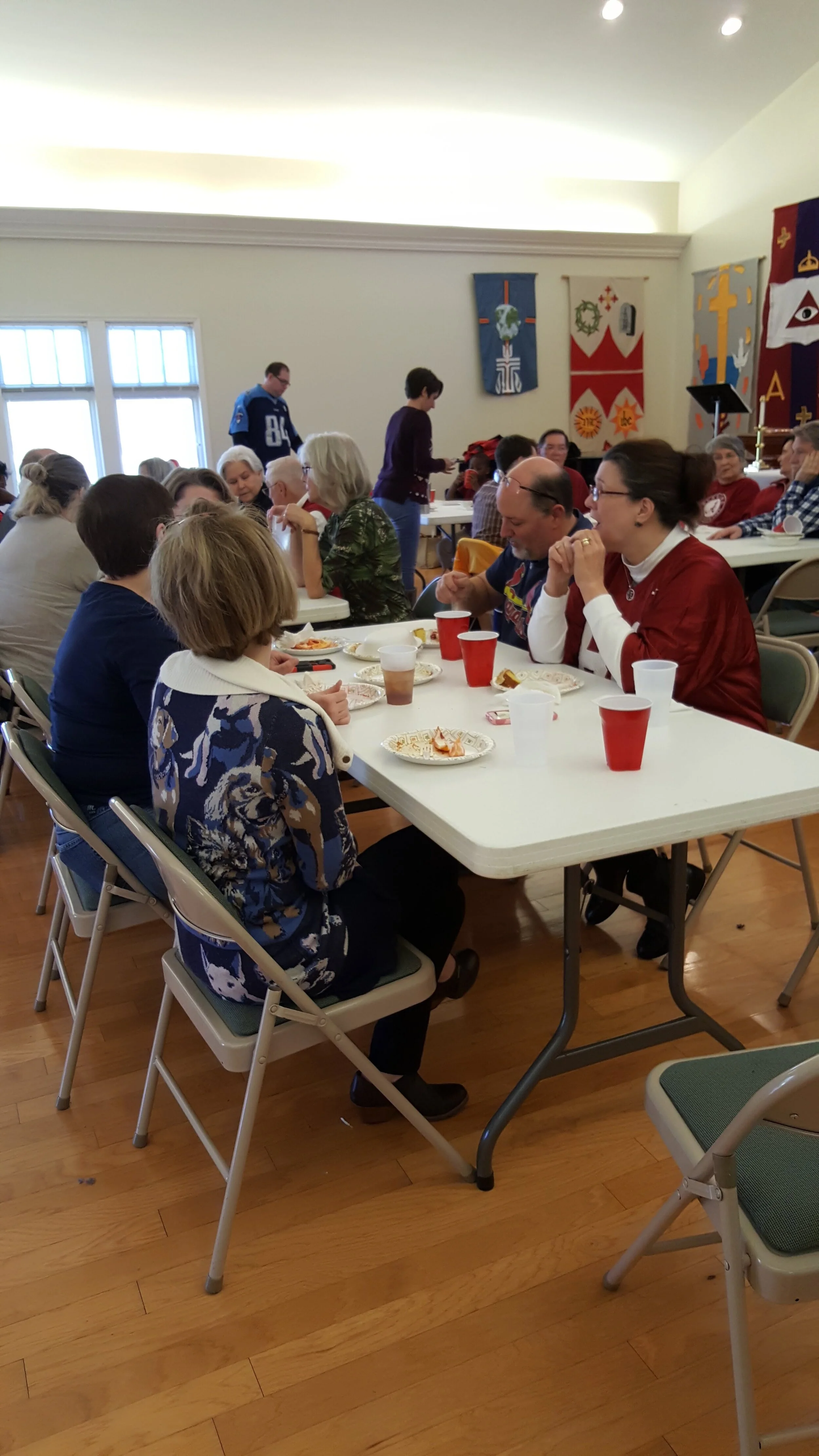 Group of people sitting at tables eating and socializing in a community hall, with decorations on the walls.