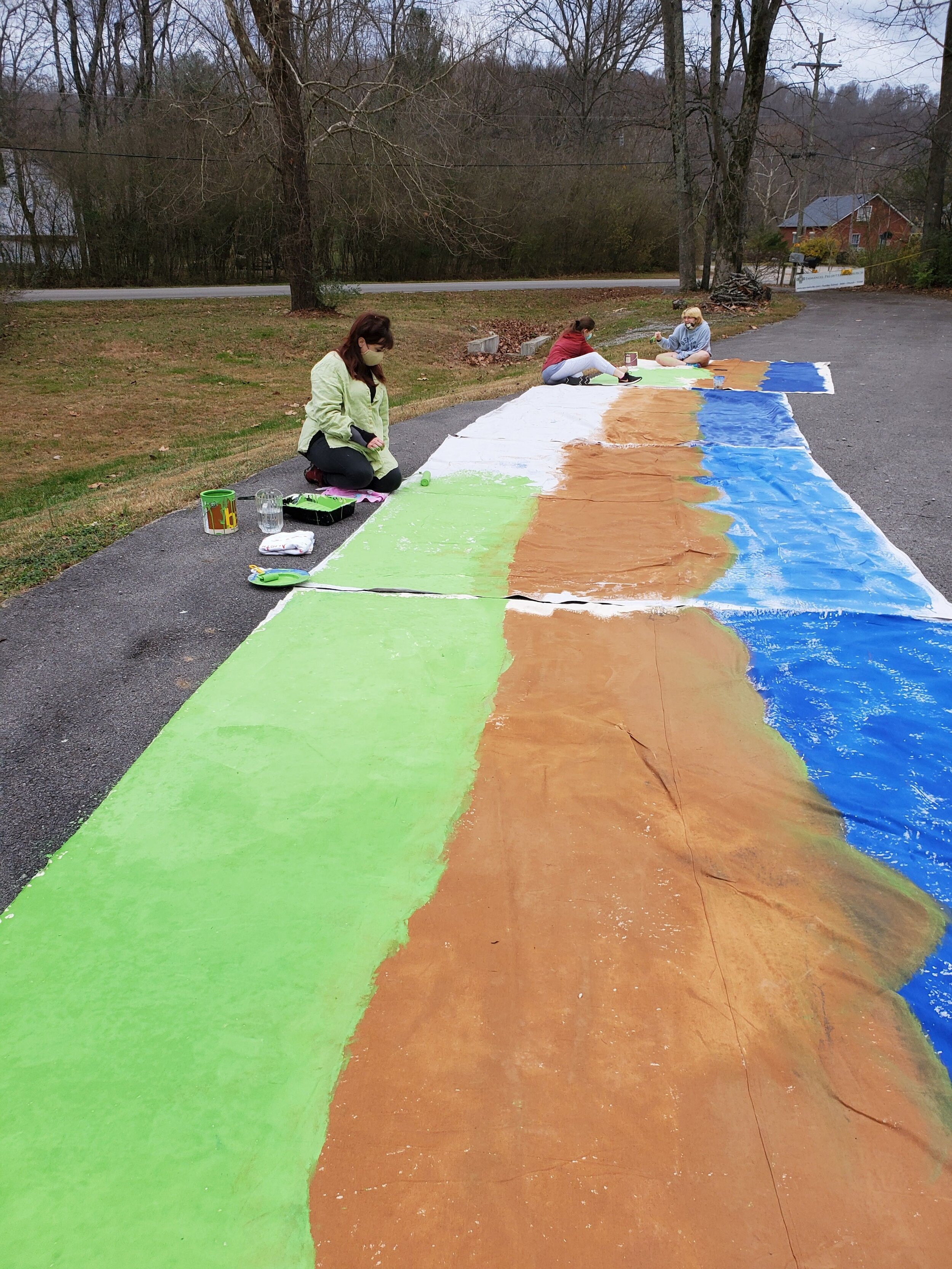 Three people painting a large outdoor mural on a road, featuring green, brown, and blue sections.