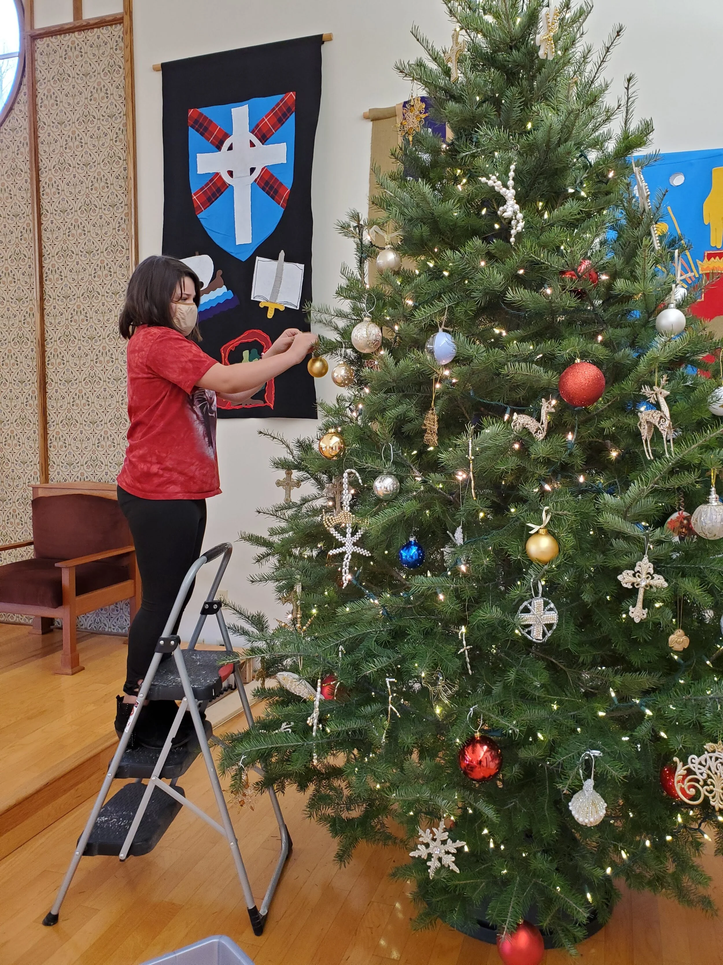 Person decorating Christmas tree with ornaments indoors while standing on a step ladder.
