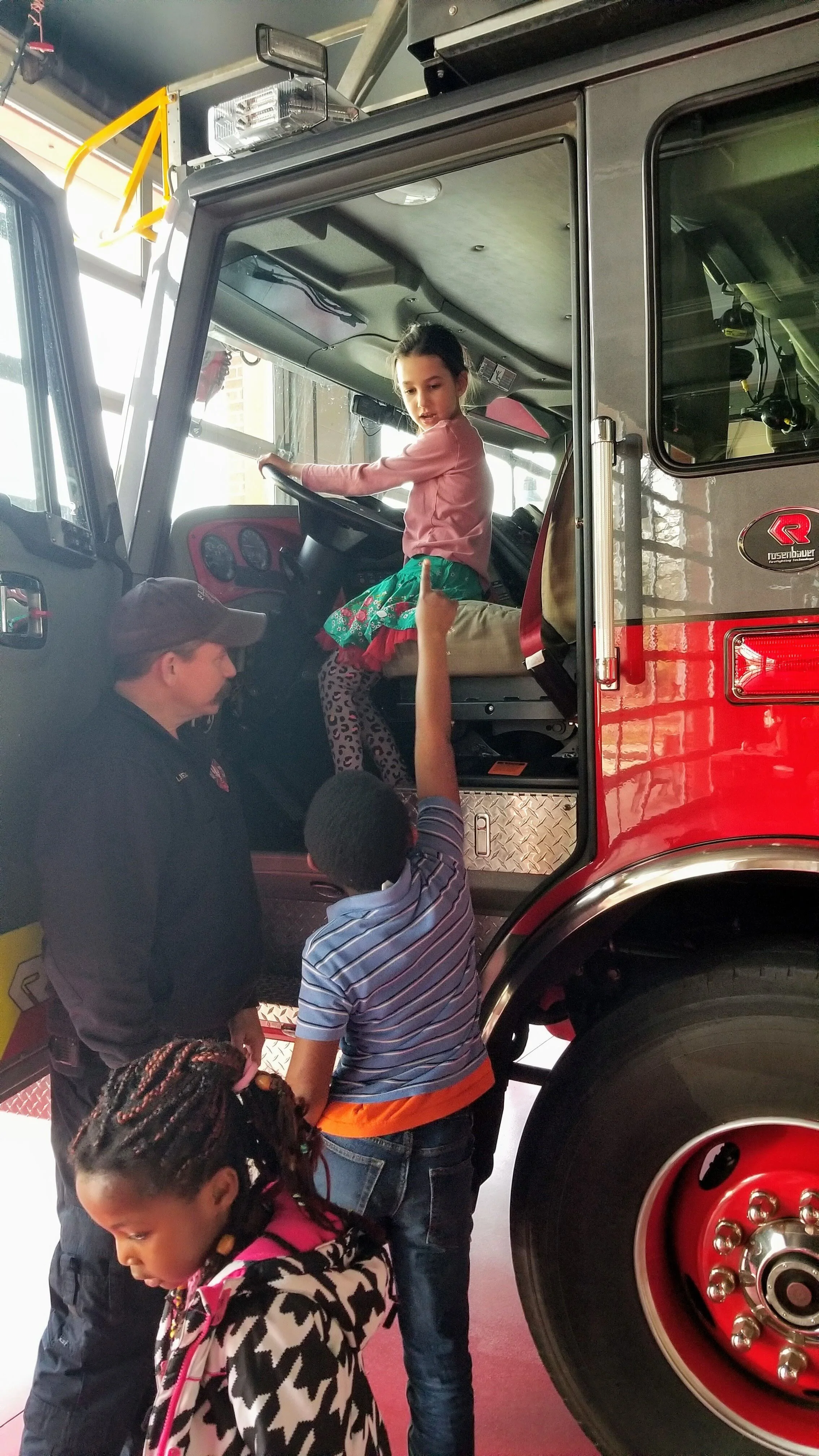 Children interacting with a firefighter in a fire truck, one child sits in the driver's seat.