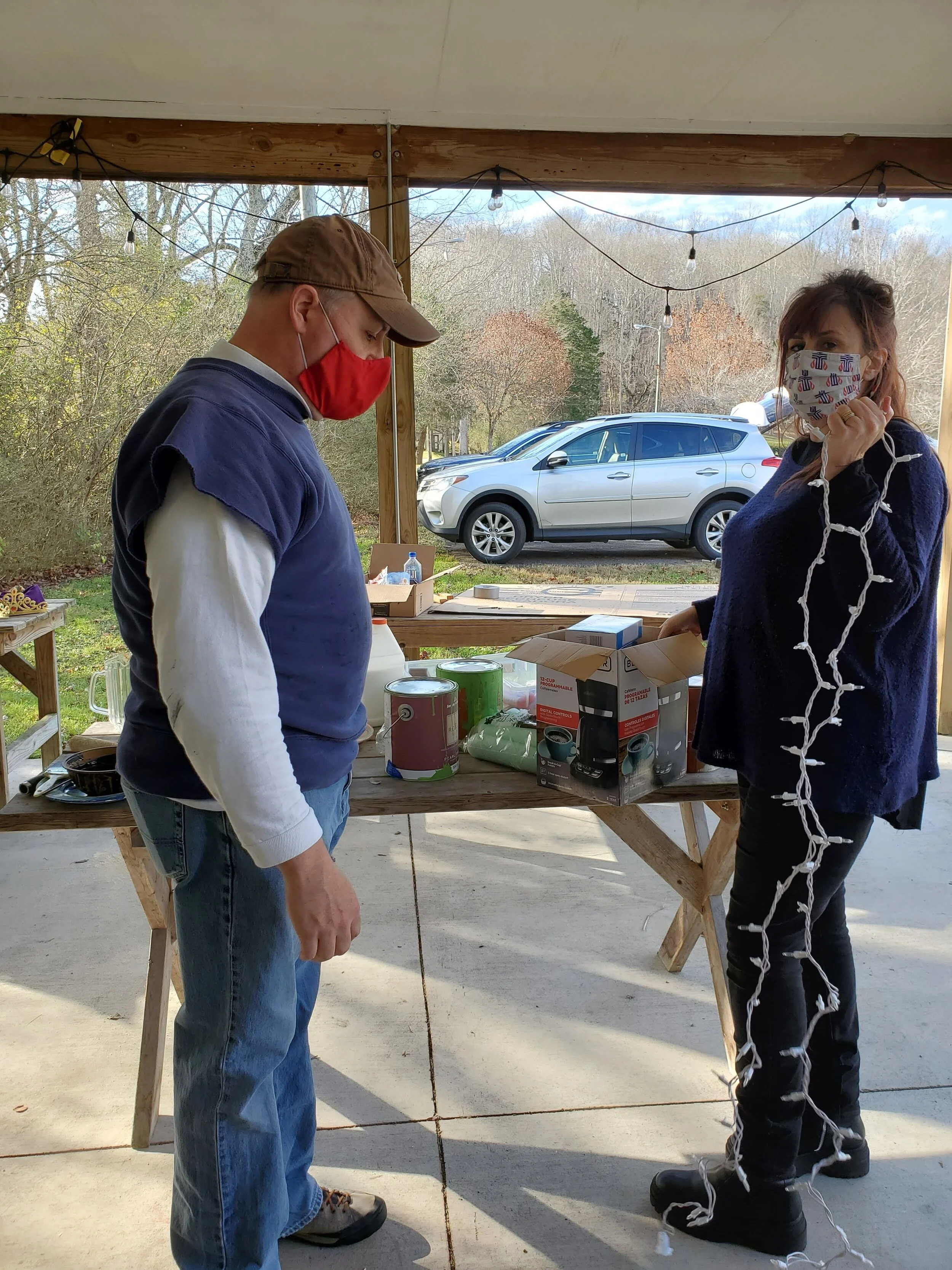 Two people wearing face masks preparing holiday decorations outdoors, with one person holding string lights and various items on a table.