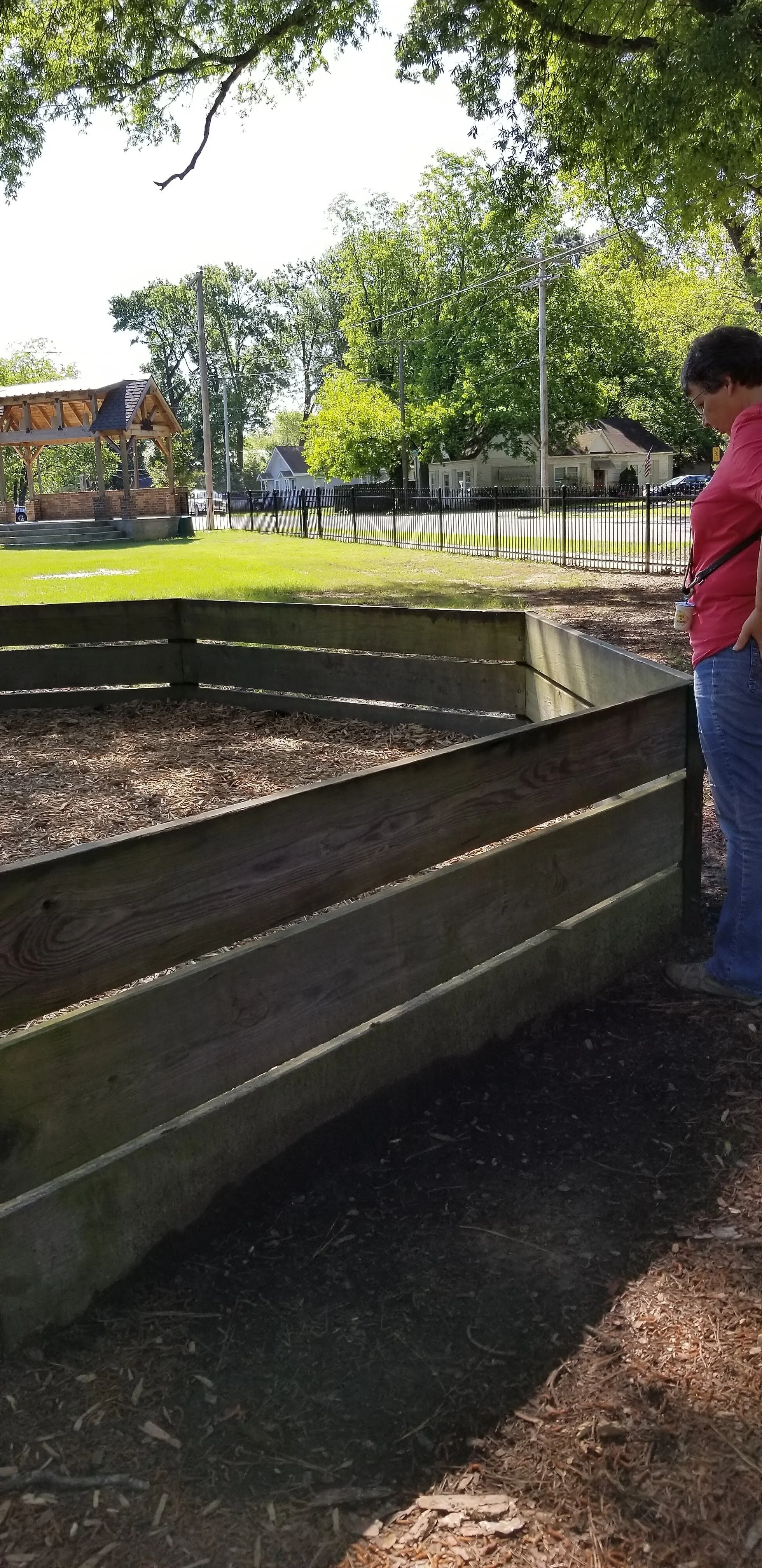 Person standing near a wooden octagonal structure, surrounded by greenery, trees, and a fenced lawn area.