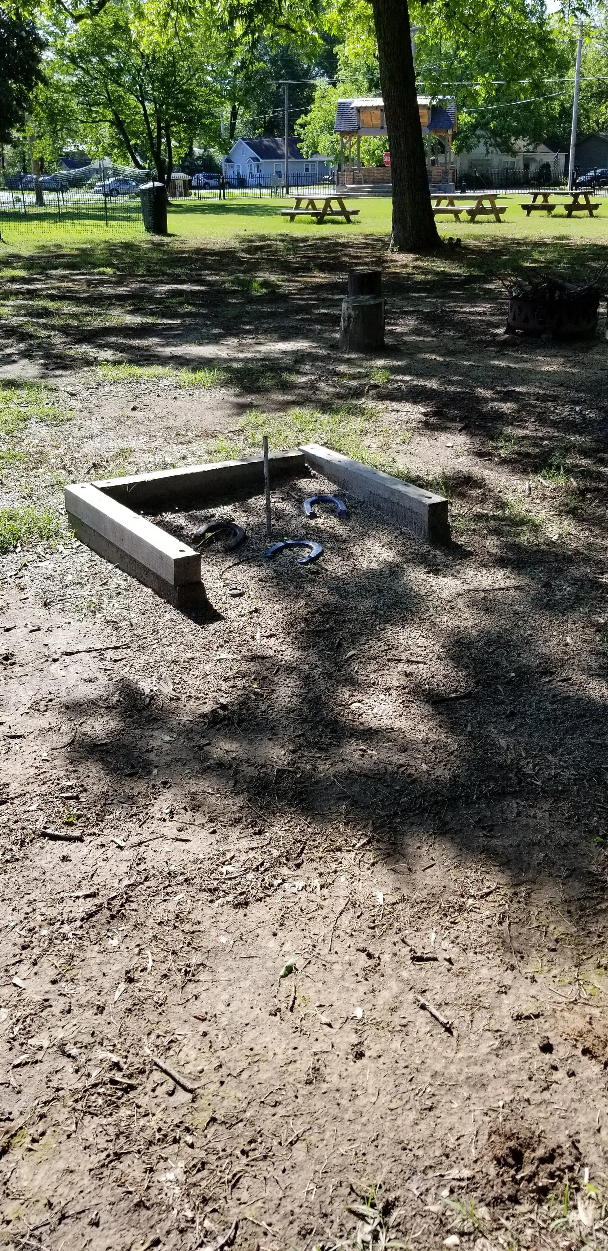 Outdoor horseshoe pit in a park setting with benches and trees.