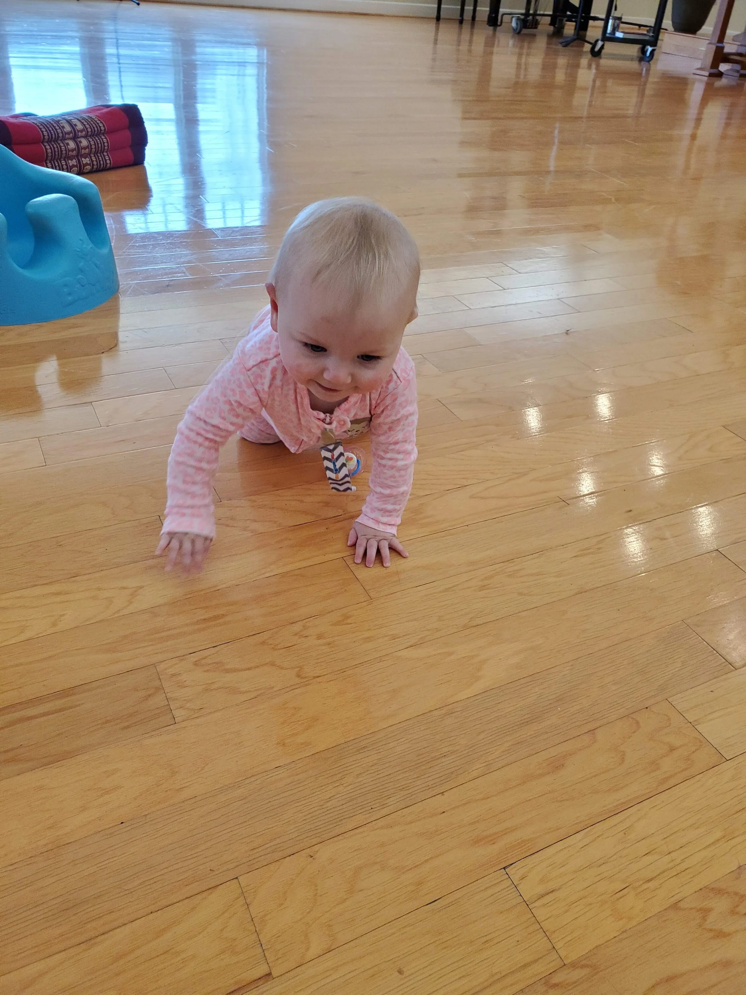 A baby in a pink outfit crawling on a wooden floor near a blue baby seat and a red cushion.