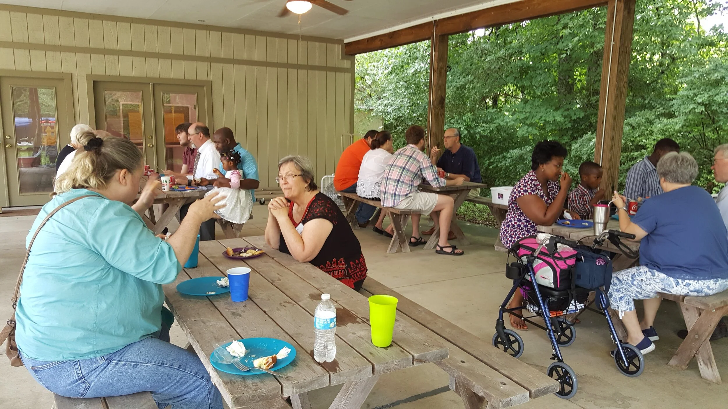 A group of people sitting at picnic tables under a covered area, eating and talking, with trees in the background.