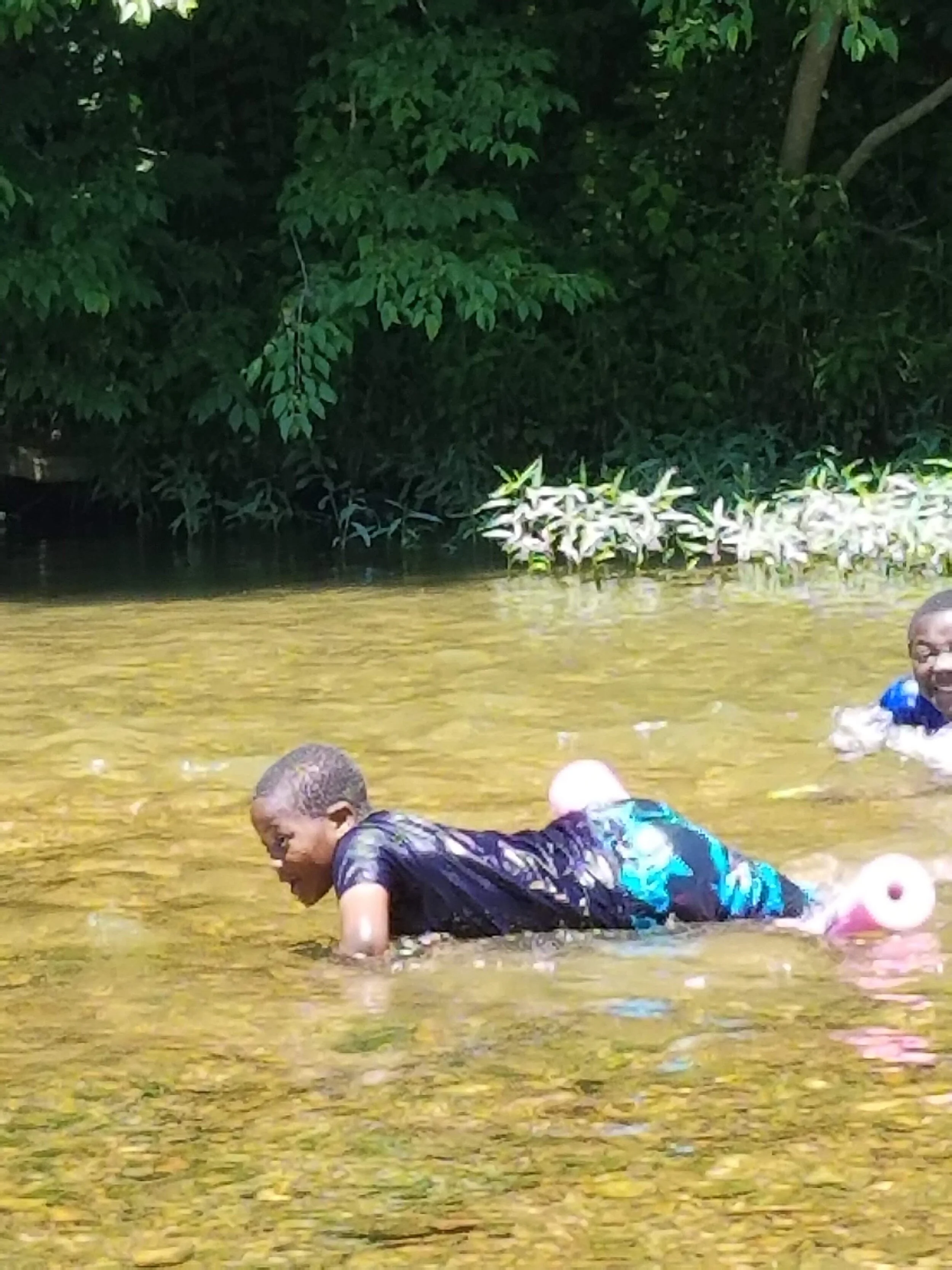 Two children playing in shallow water with trees in the background, one using a pool noodle.