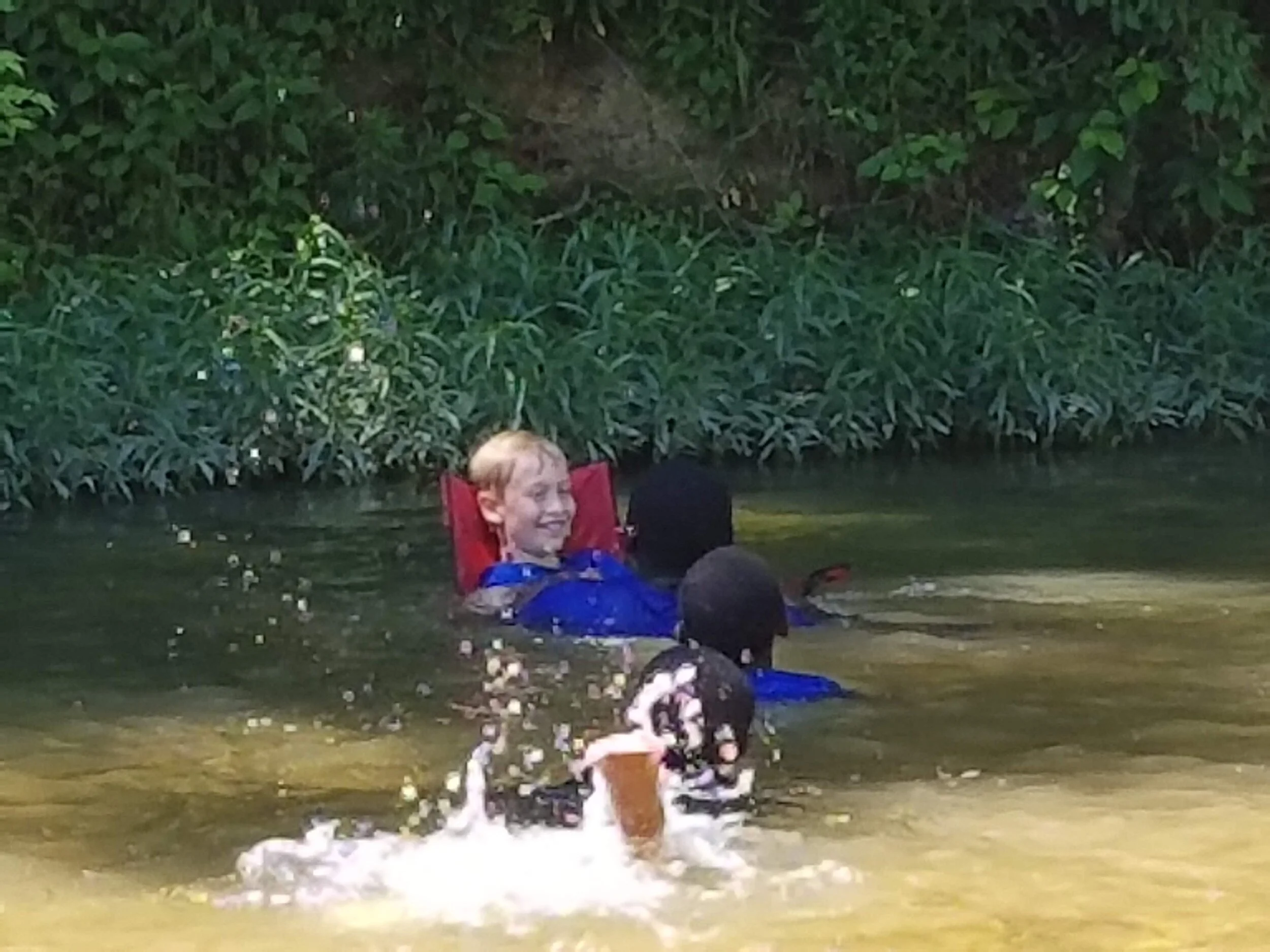 Children playing and swimming in a river wearing blue life vests, with greenery in the background.