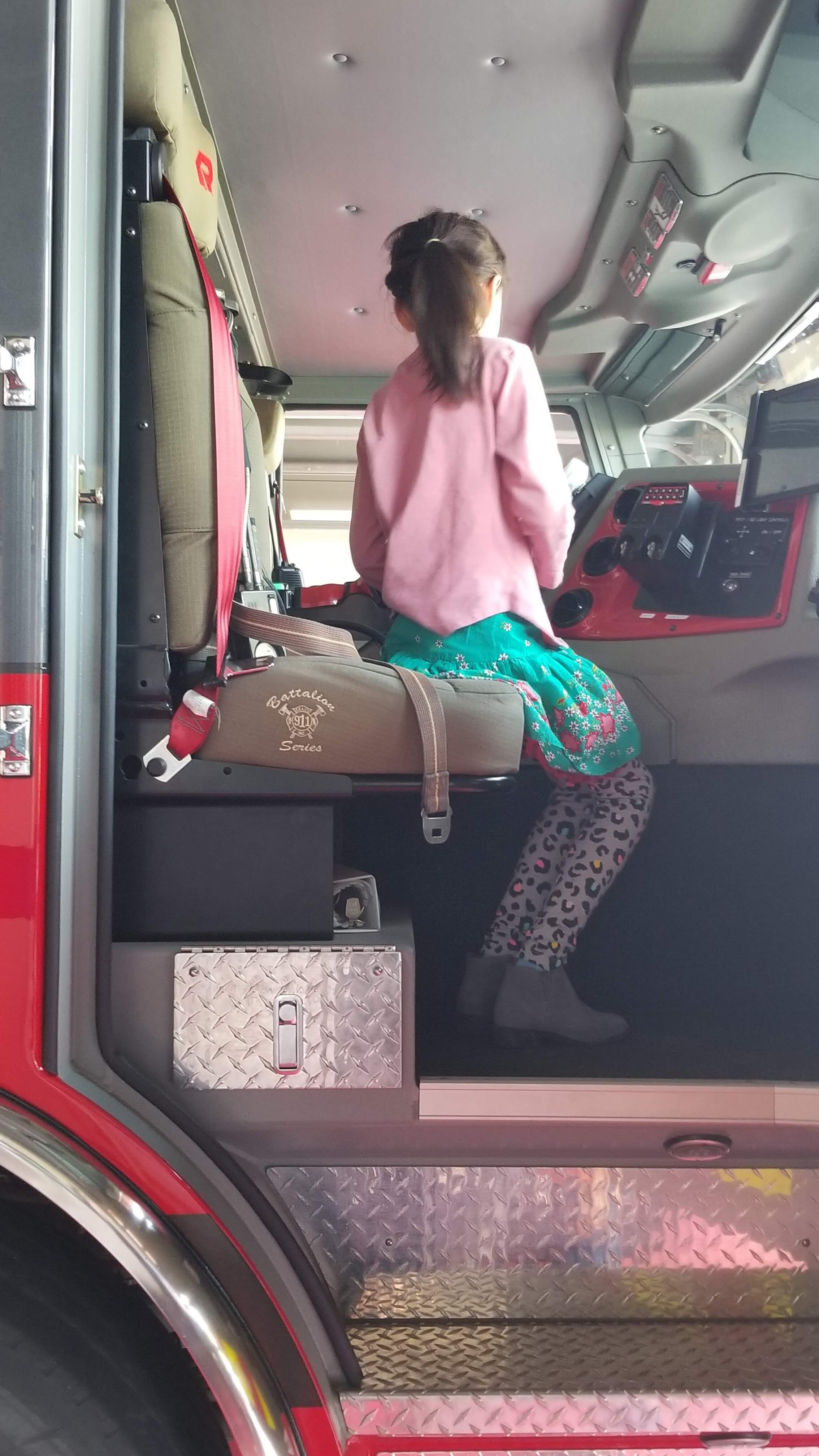 Child sitting in the driver's seat of a fire truck, viewed from the side, with interior details visible.