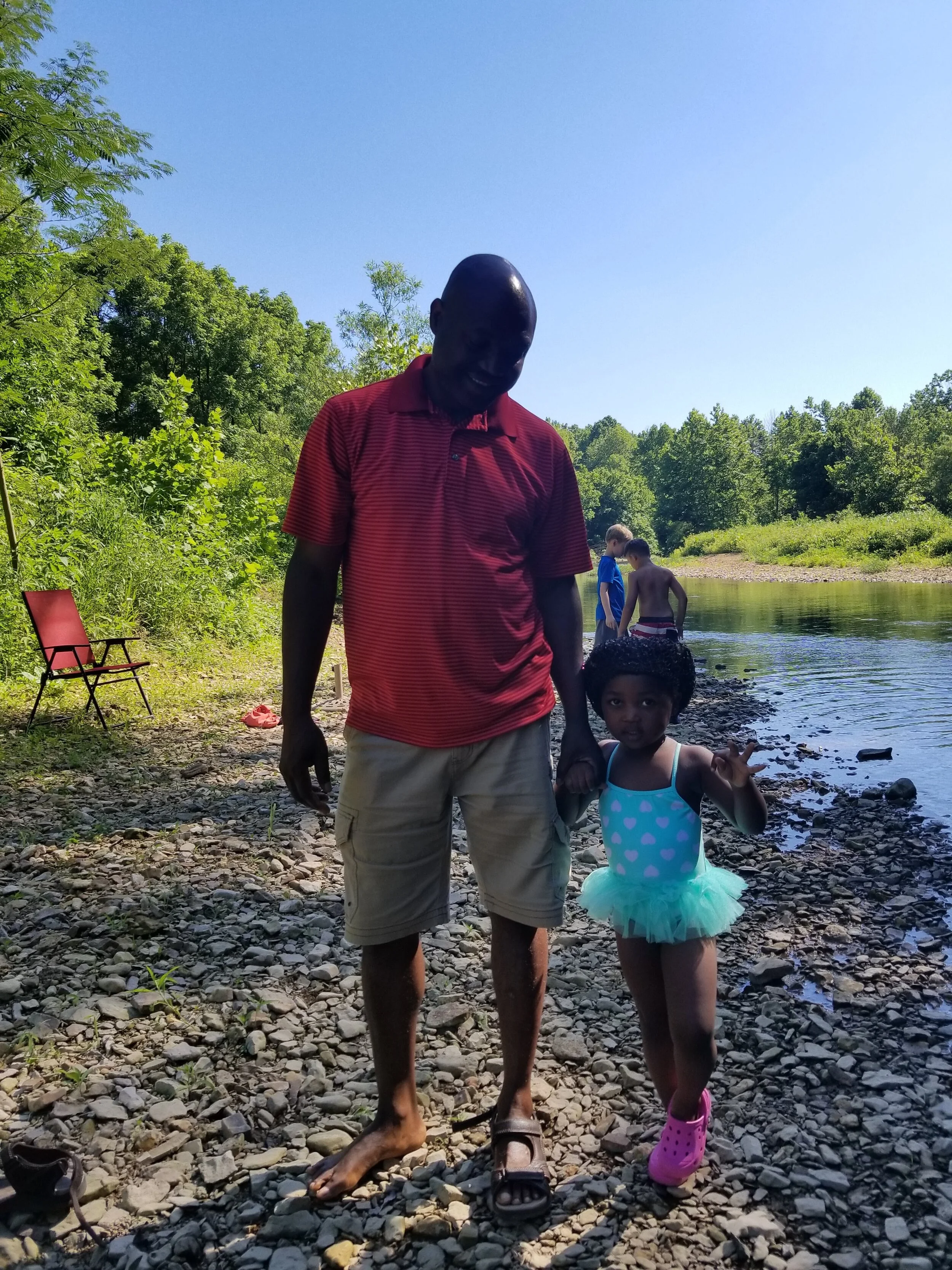Man in red shirt and khaki shorts with young girl in blue swimsuit and pink shoes standing by a rocky riverside, surrounded by green trees under a clear blue sky.