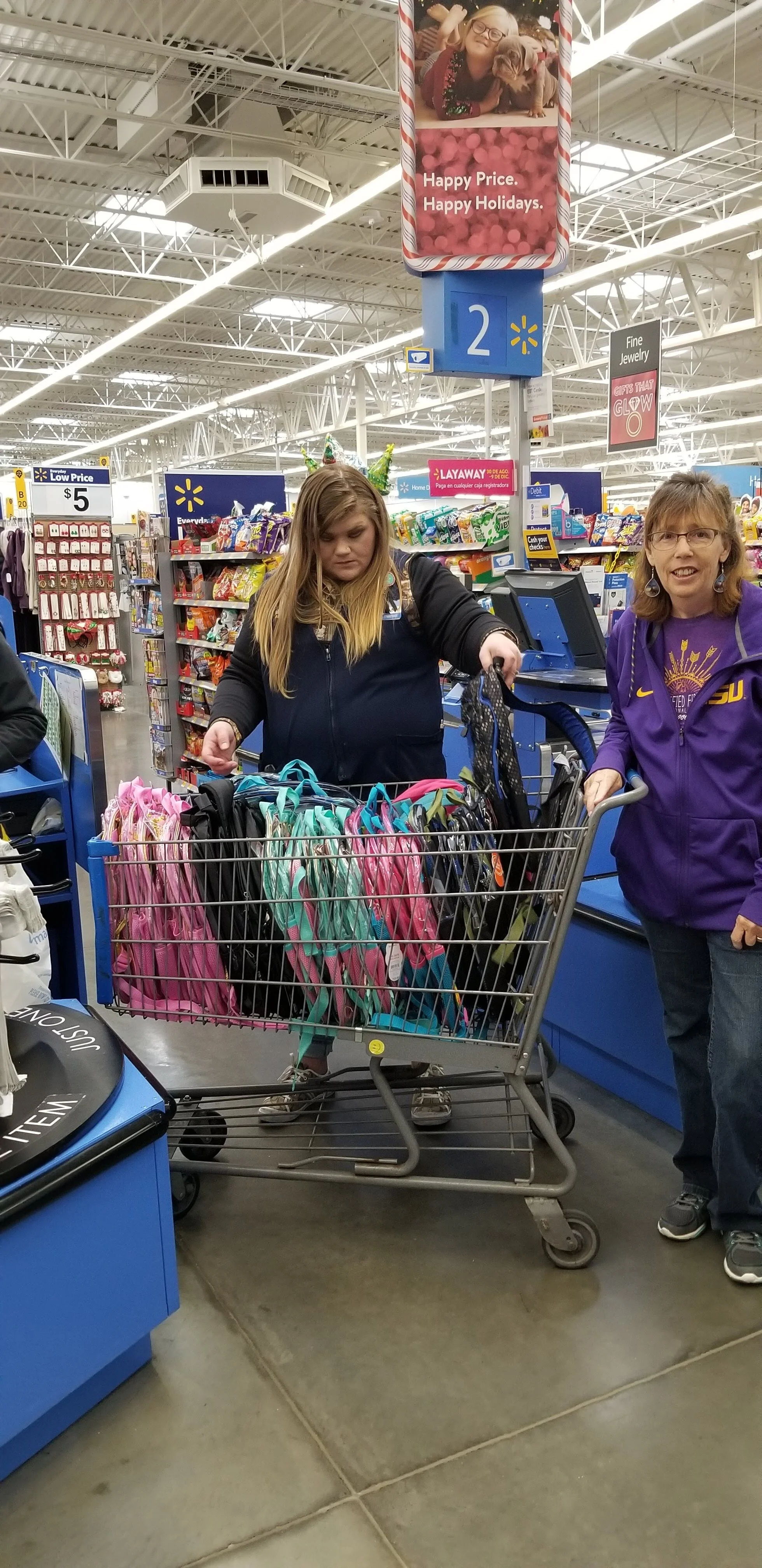 Customers at a retail store checkout with a cart full of colorful backpacks, under a festive holiday sign.