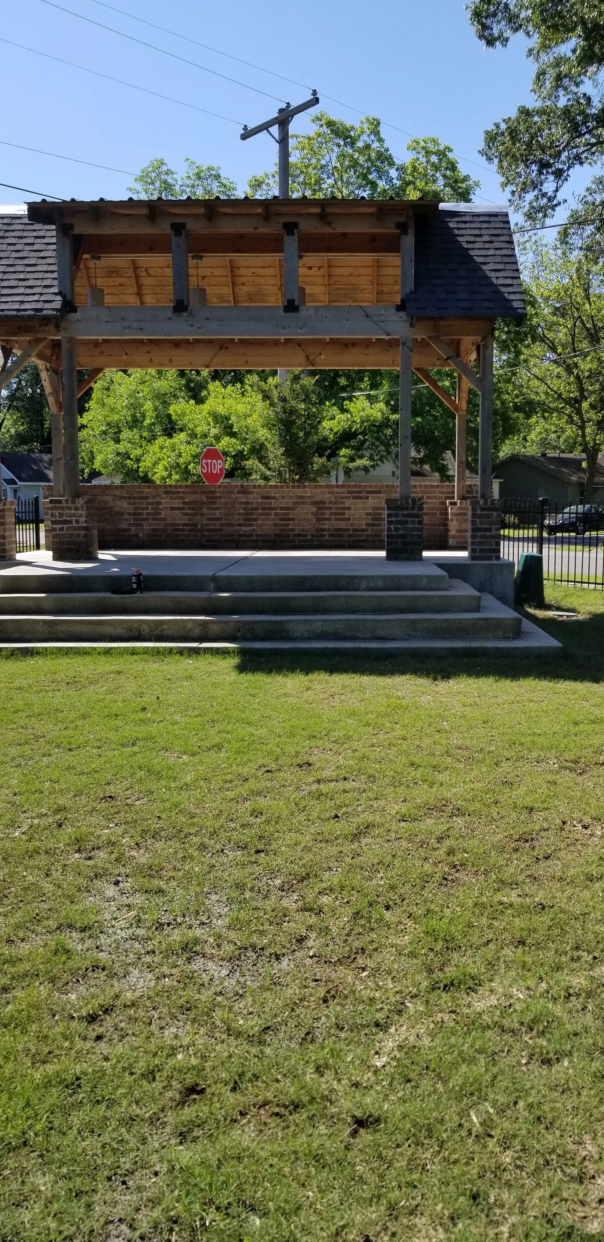 Outdoor pavilion with brick base and wooden roof, concrete steps leading to platform, stop sign in the background, grass lawn in foreground.