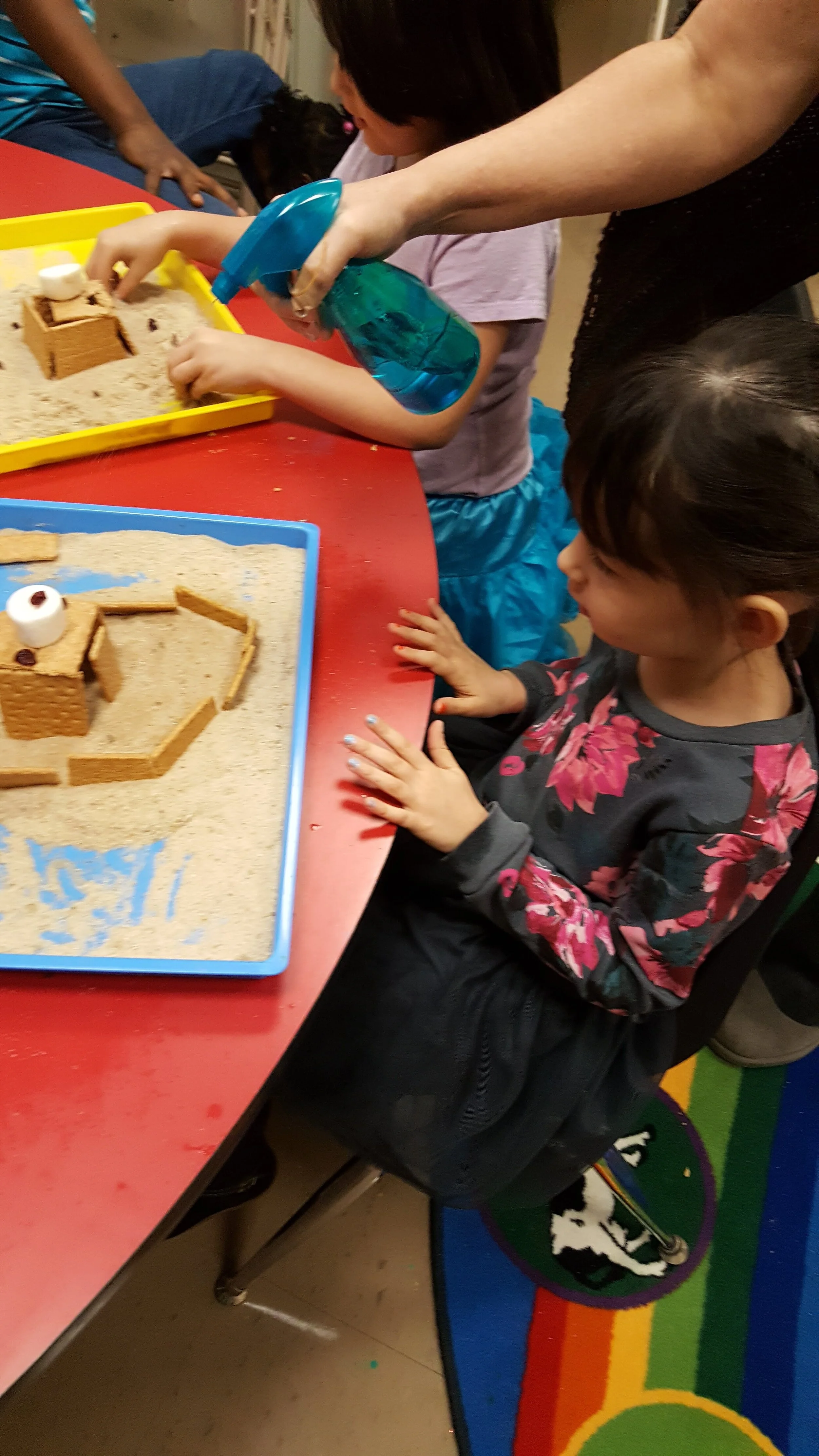 Children at a table building structures with sand and graham crackers, with one child using a spray bottle.
