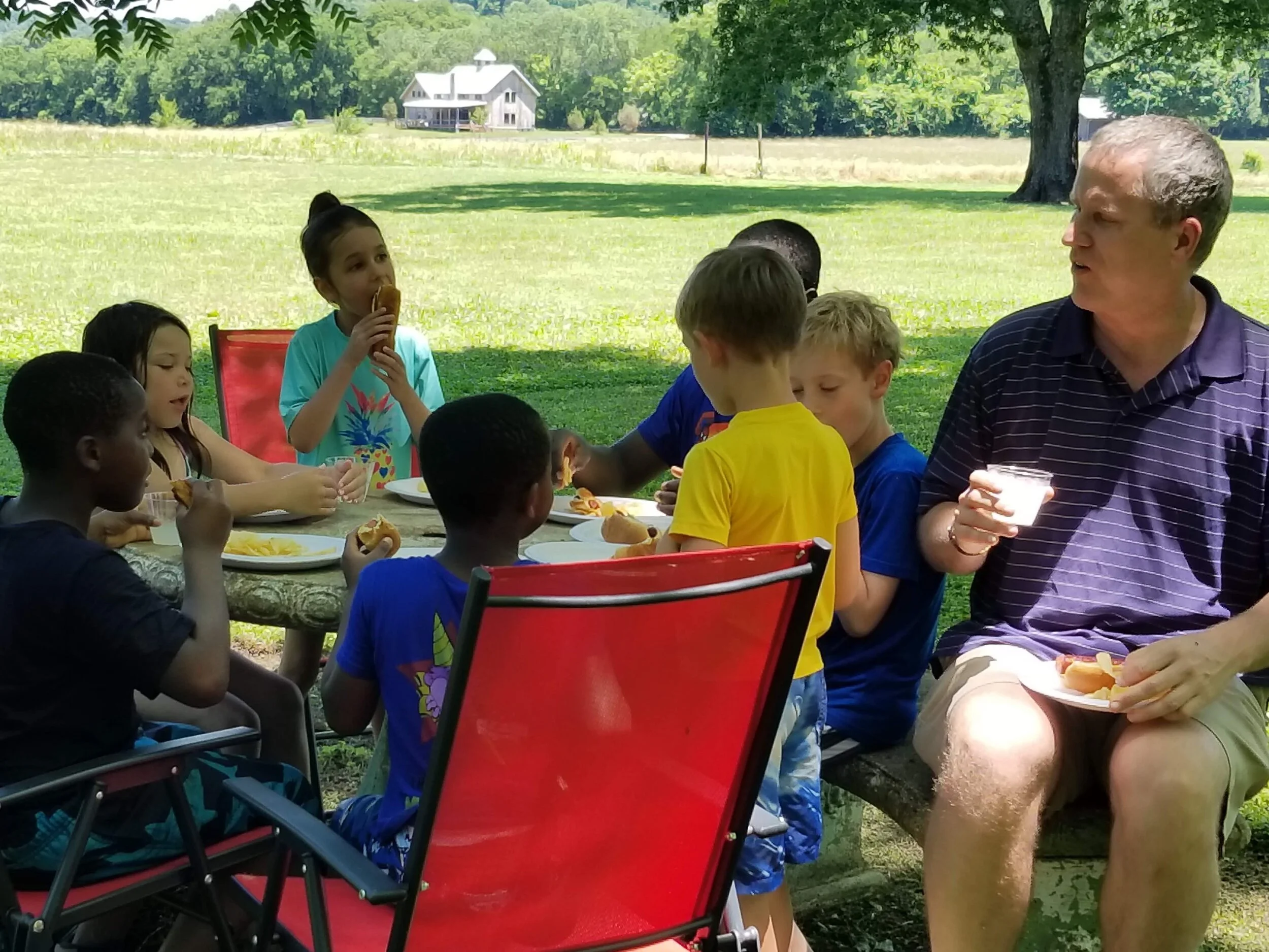 Group of kids and an adult eating hot dogs and fries at an outdoor picnic table in a park setting.