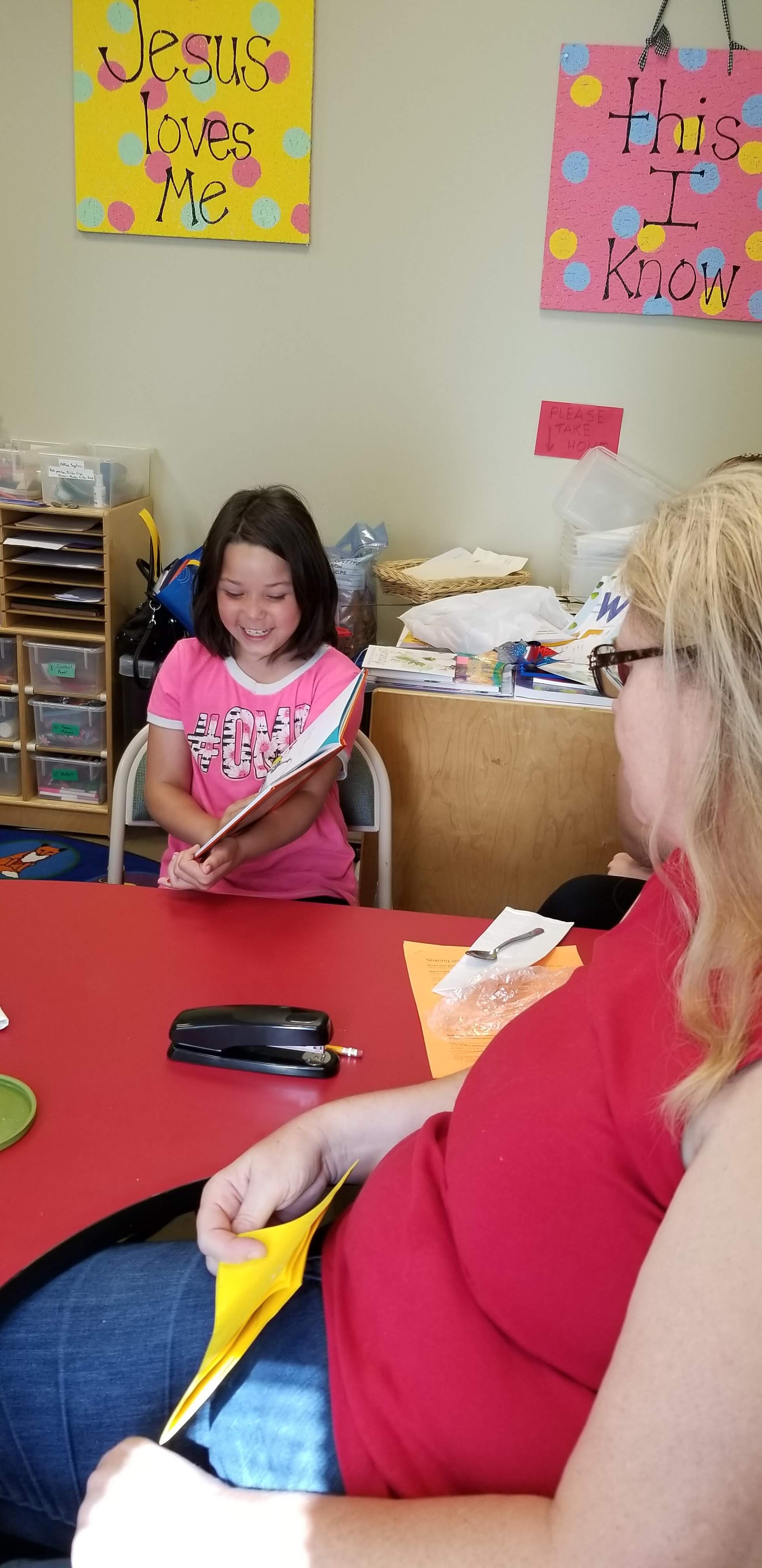 Child reading a book and smiling in a classroom setting, with an adult seated nearby. A yellow sign on the wall reads "Jesus loves me" and a pink sign says "this I know." Classroom supplies are visible in the background.
