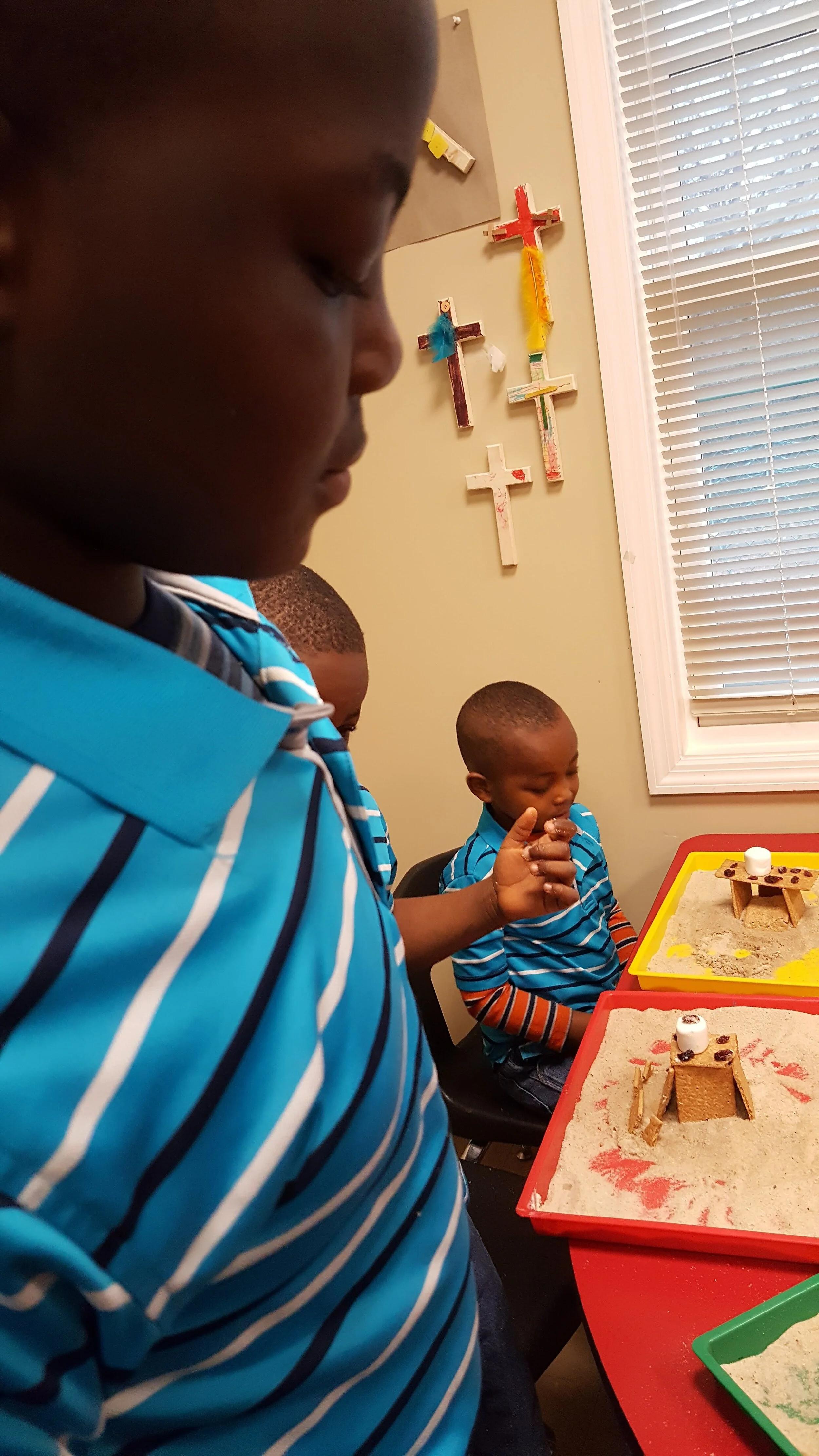 Children wearing striped shirts working on creative crafts with sand and graham crackers, surrounded by handmade crosses on the wall.