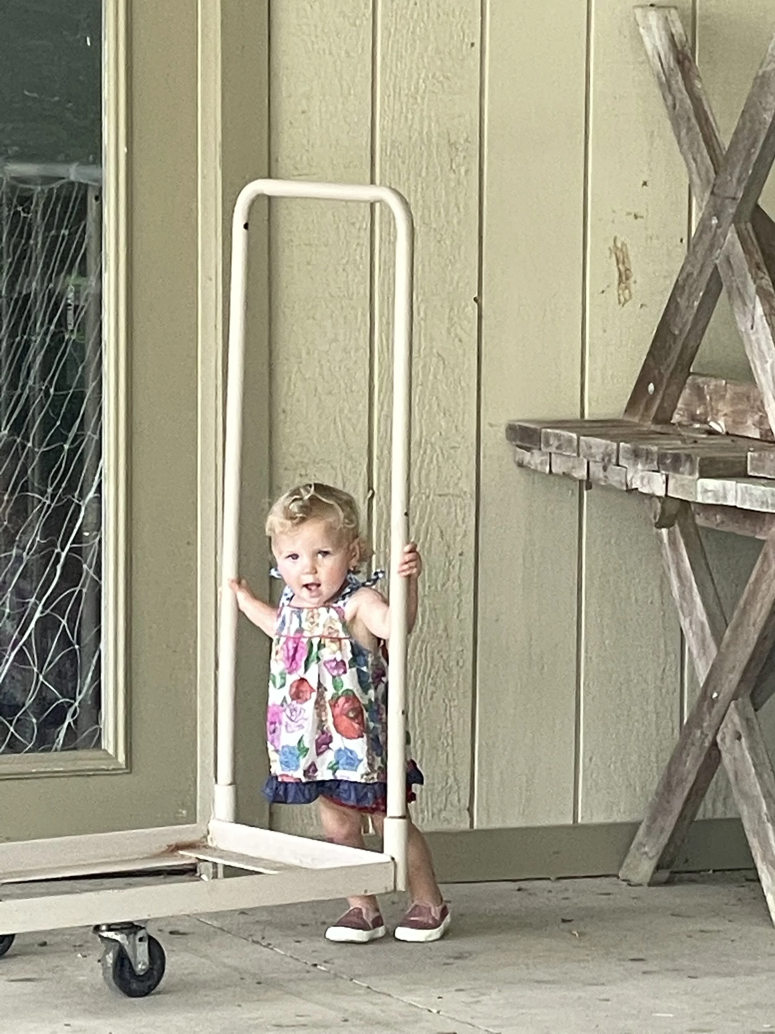 A toddler in a floral dress standing beside a metal hand truck on a patio, with a wooden wall and plant stand in the background.