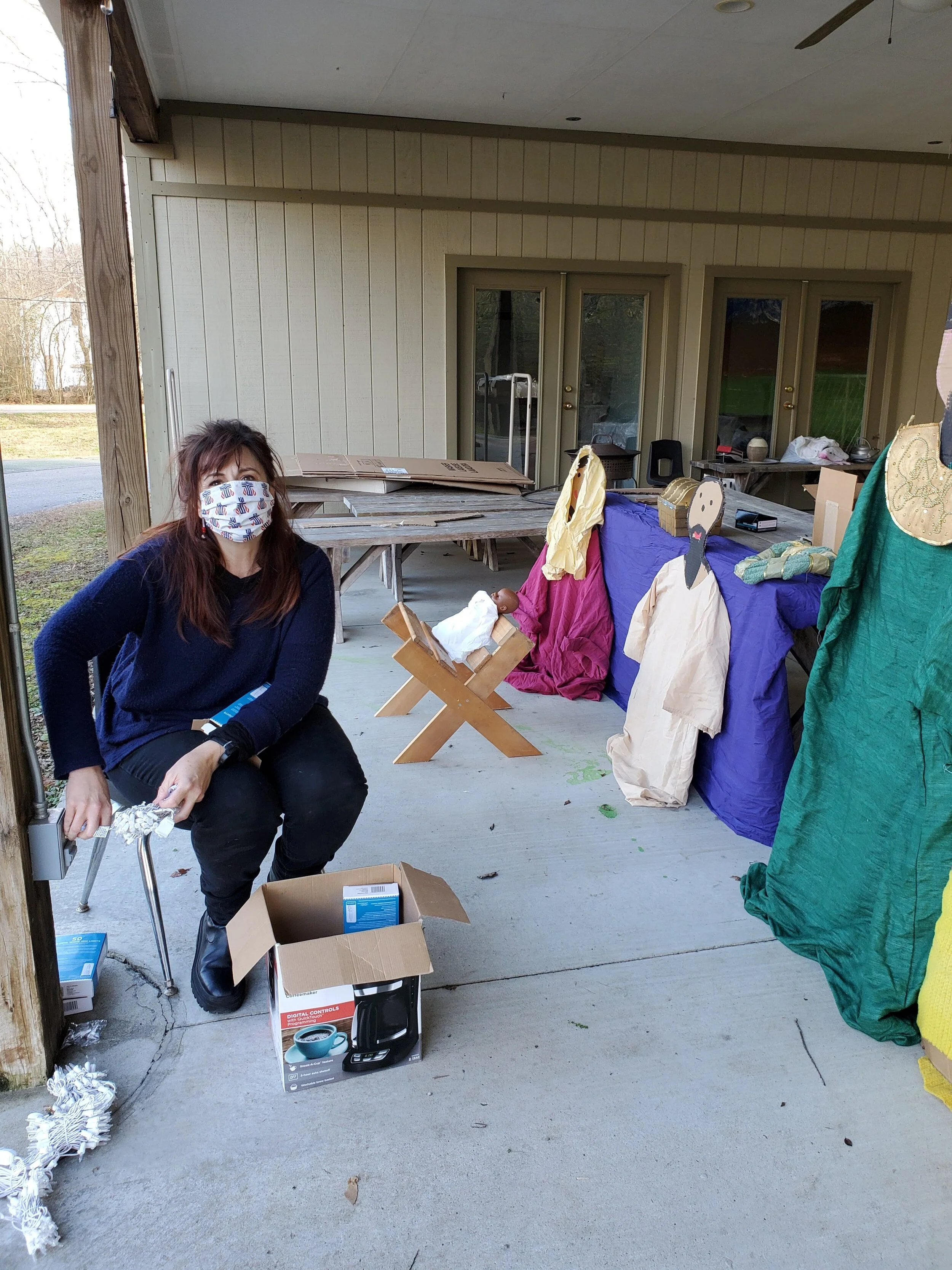 Woman wearing a face mask sitting on a chair next to an open box, with a nativity set and wrapped objects on a covered porch.