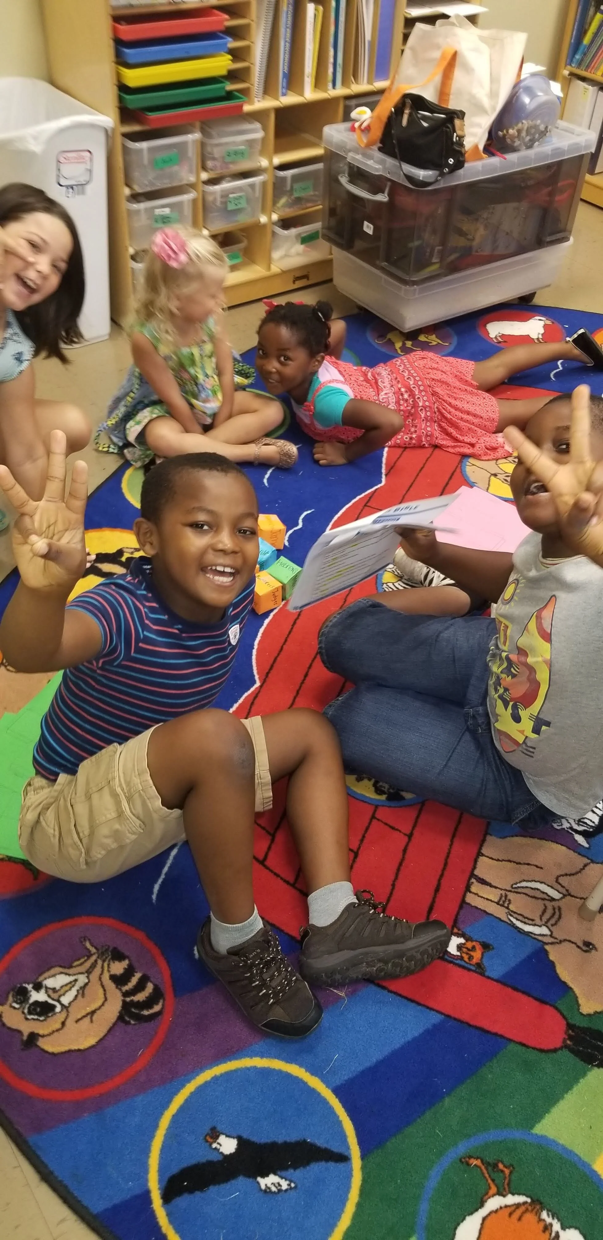 Children sitting on an animal-themed carpet in a classroom, smiling and showing peace signs.
