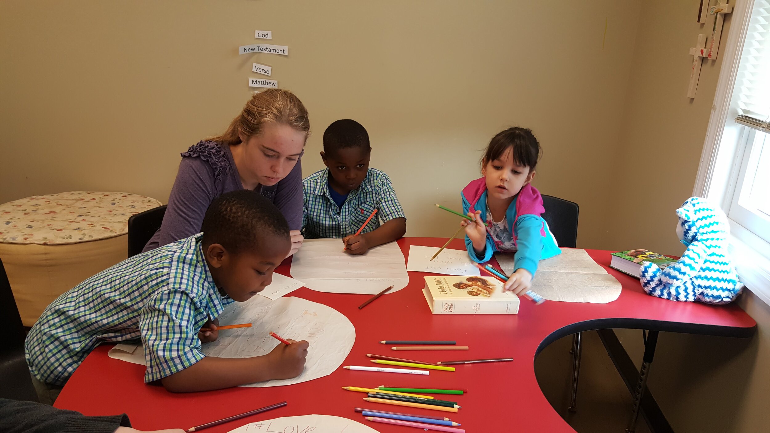 Children and a teacher drawing with colored pencils on a red table in a classroom.
