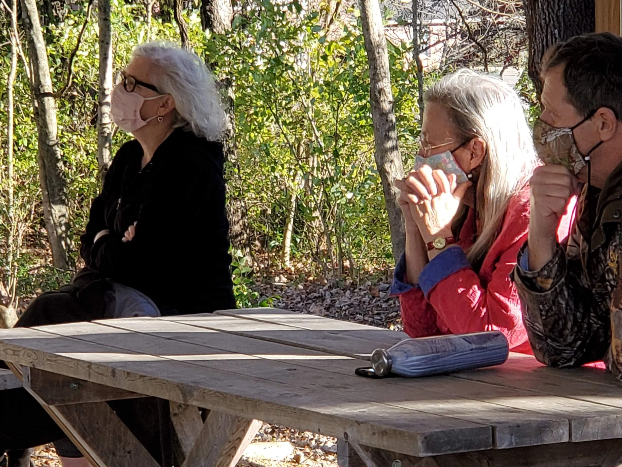 Three people sitting at a picnic table outdoors, wearing face masks.
