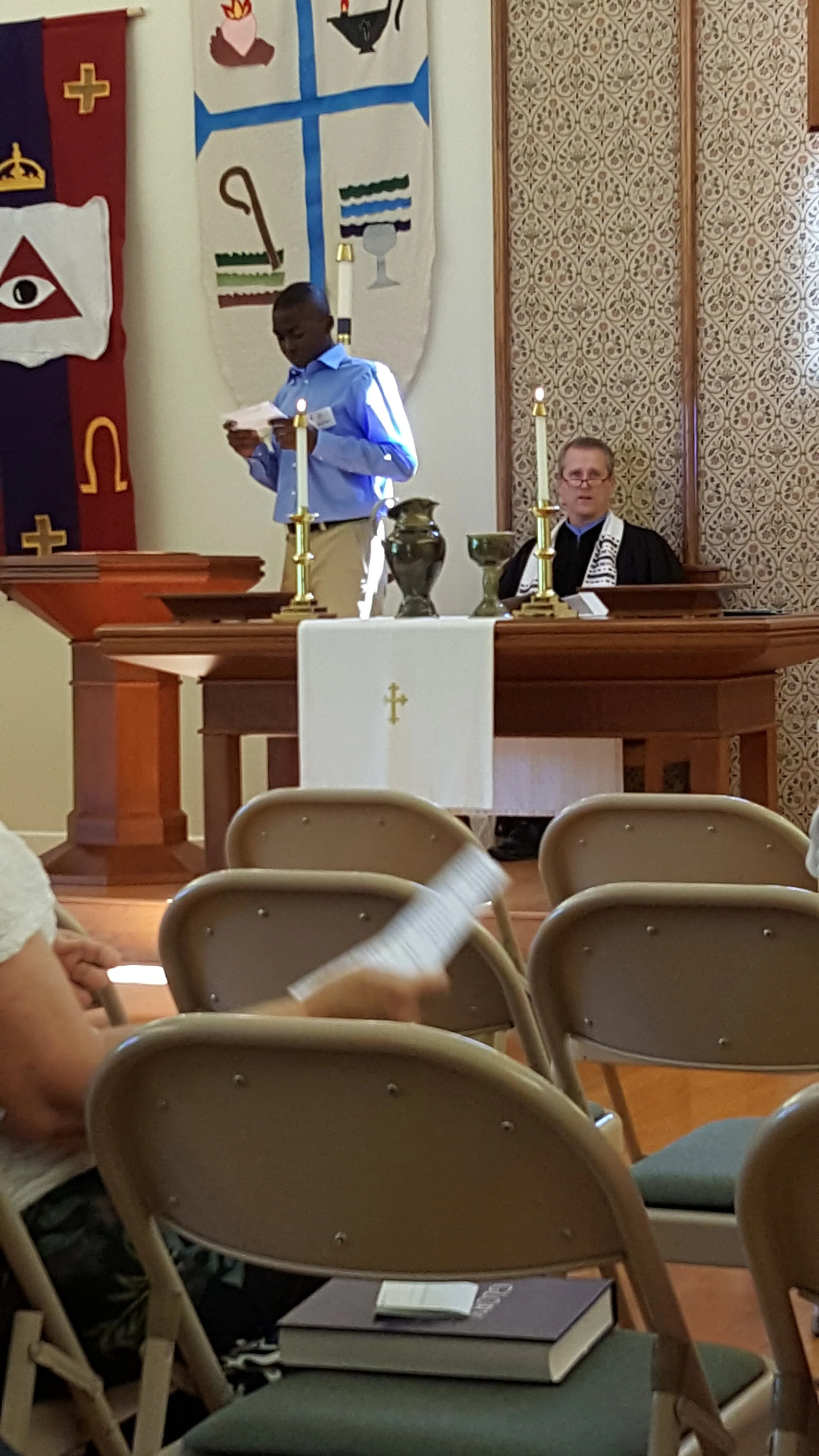 A church interior showing a person standing and reading from a paper, with another seated at a table with religious symbols, candlesticks, and decorative banners in the background.