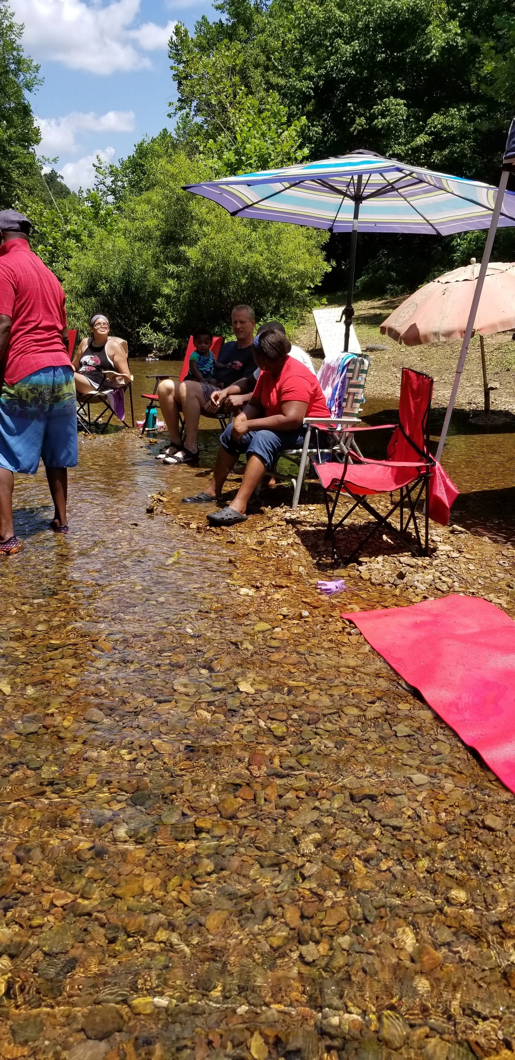 Group of people sitting in a shallow stream with chairs and umbrellas, surrounded by greenery and clear water; some have their feet in the water, with towels and blankets nearby.