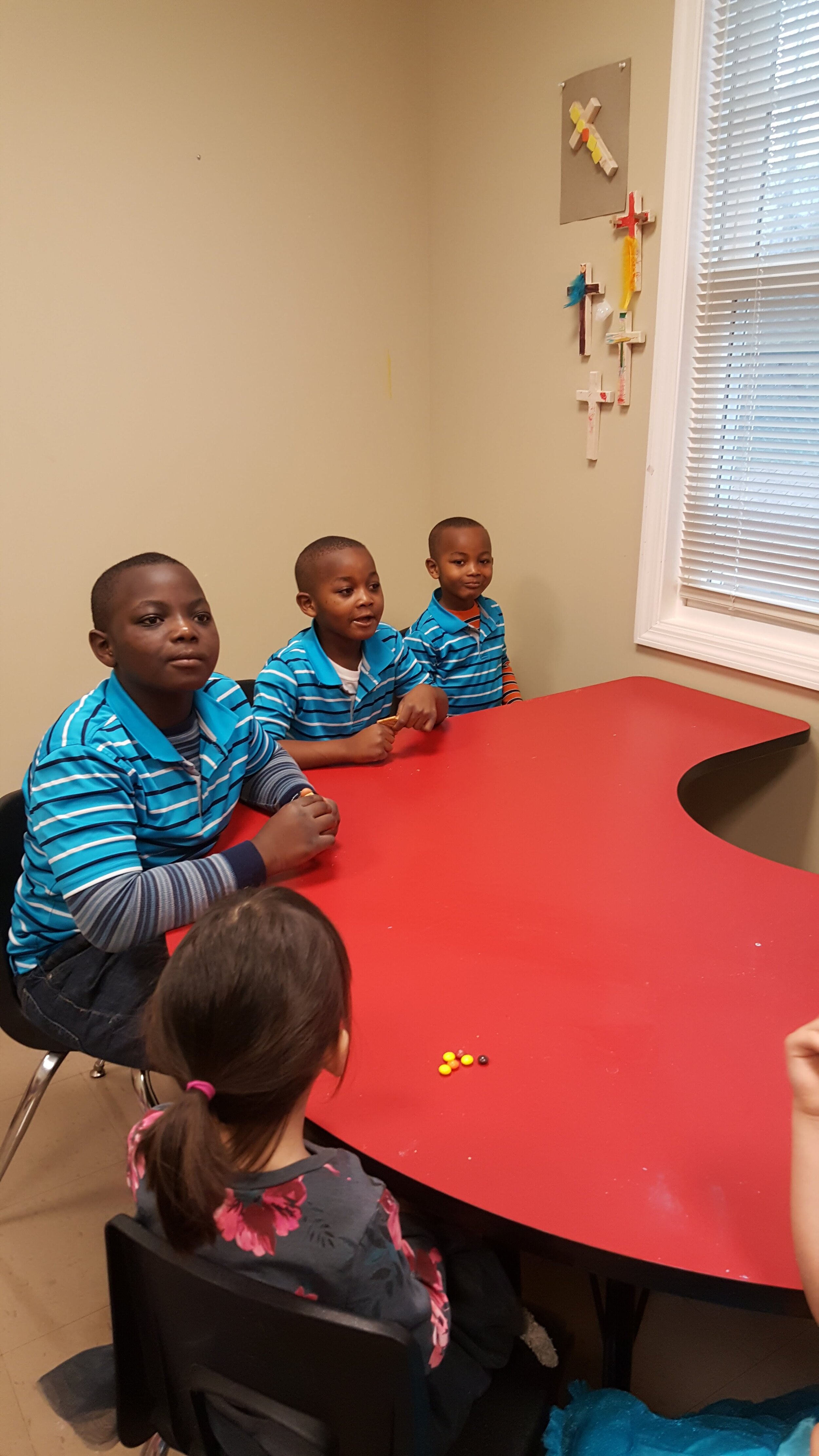 Children at a red table in a classroom, wearing striped shirts.