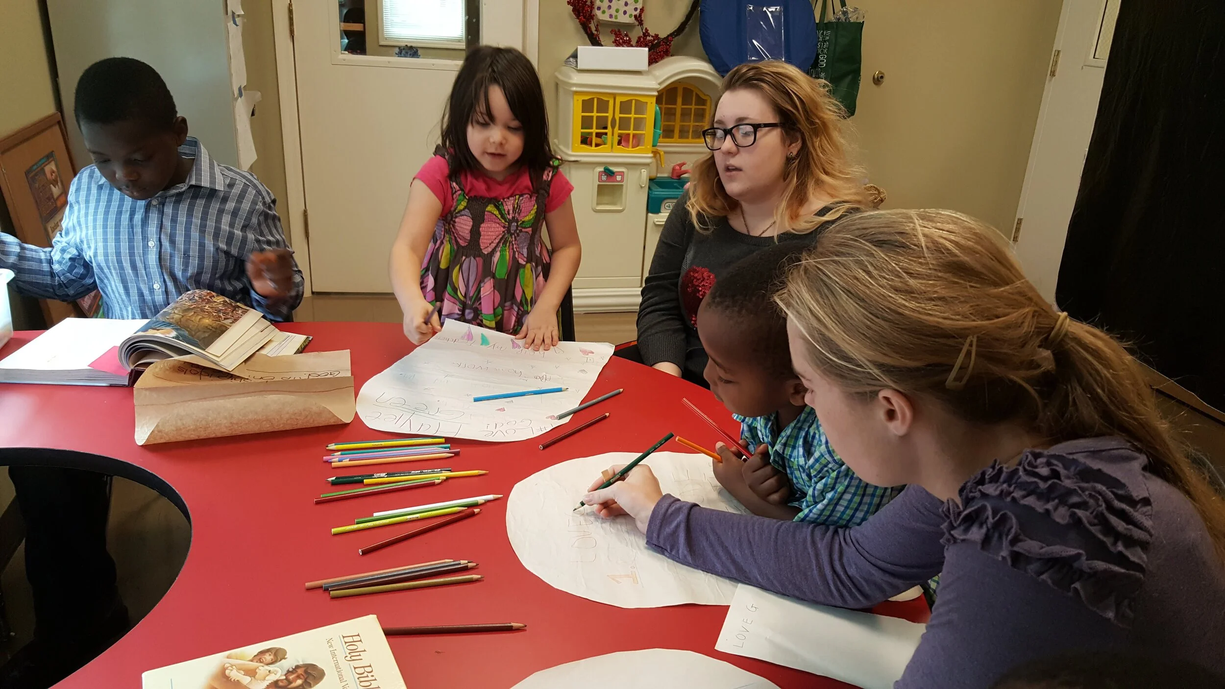 A group of children and an adult engaged in an arts and crafts activity around a red table, with colored pencils and paper. One child is holding a marker near a large piece of paper. A book is visible in the foreground.