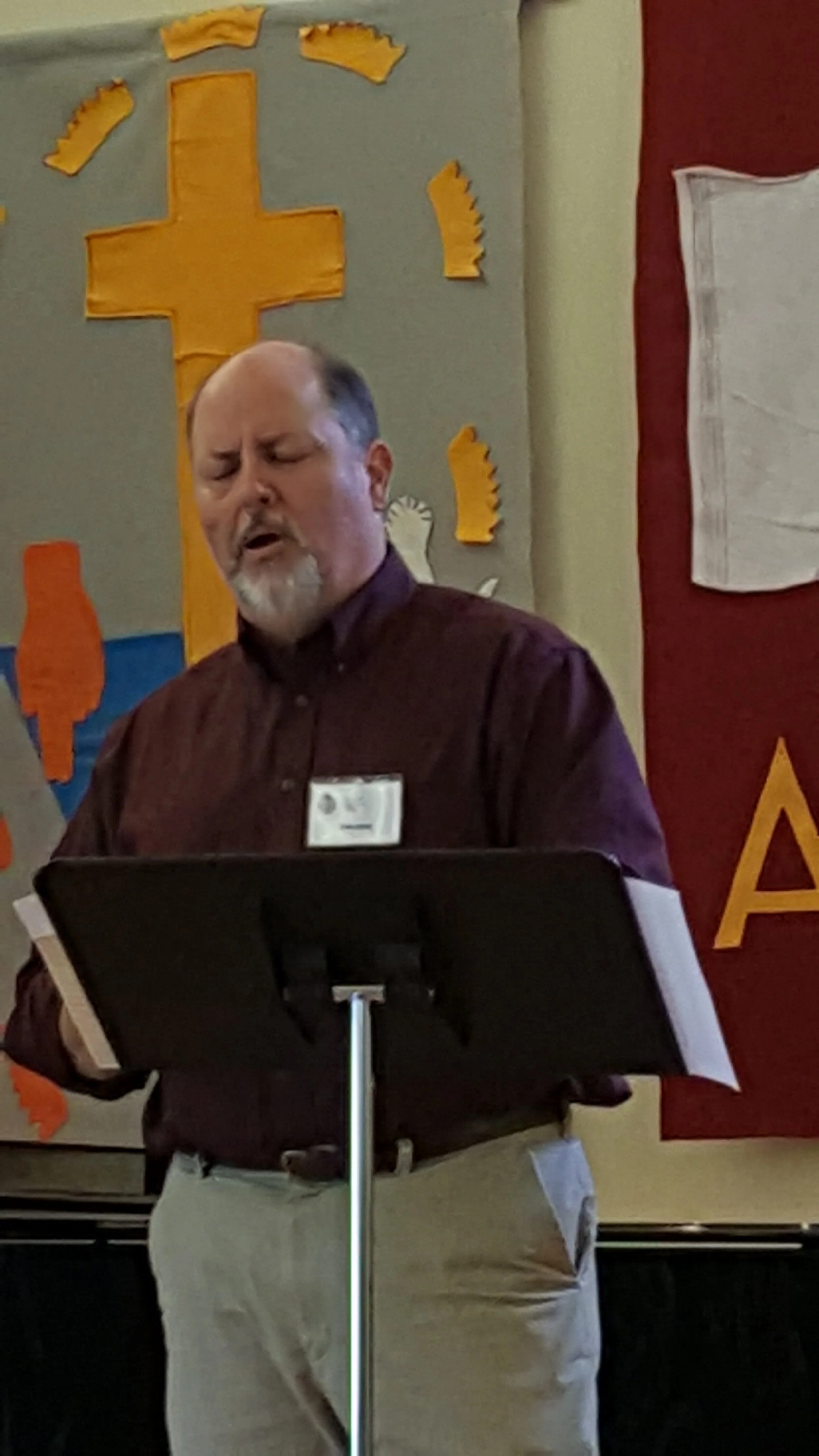 Man speaking with a stand, beside a cross and flag banner in a room.