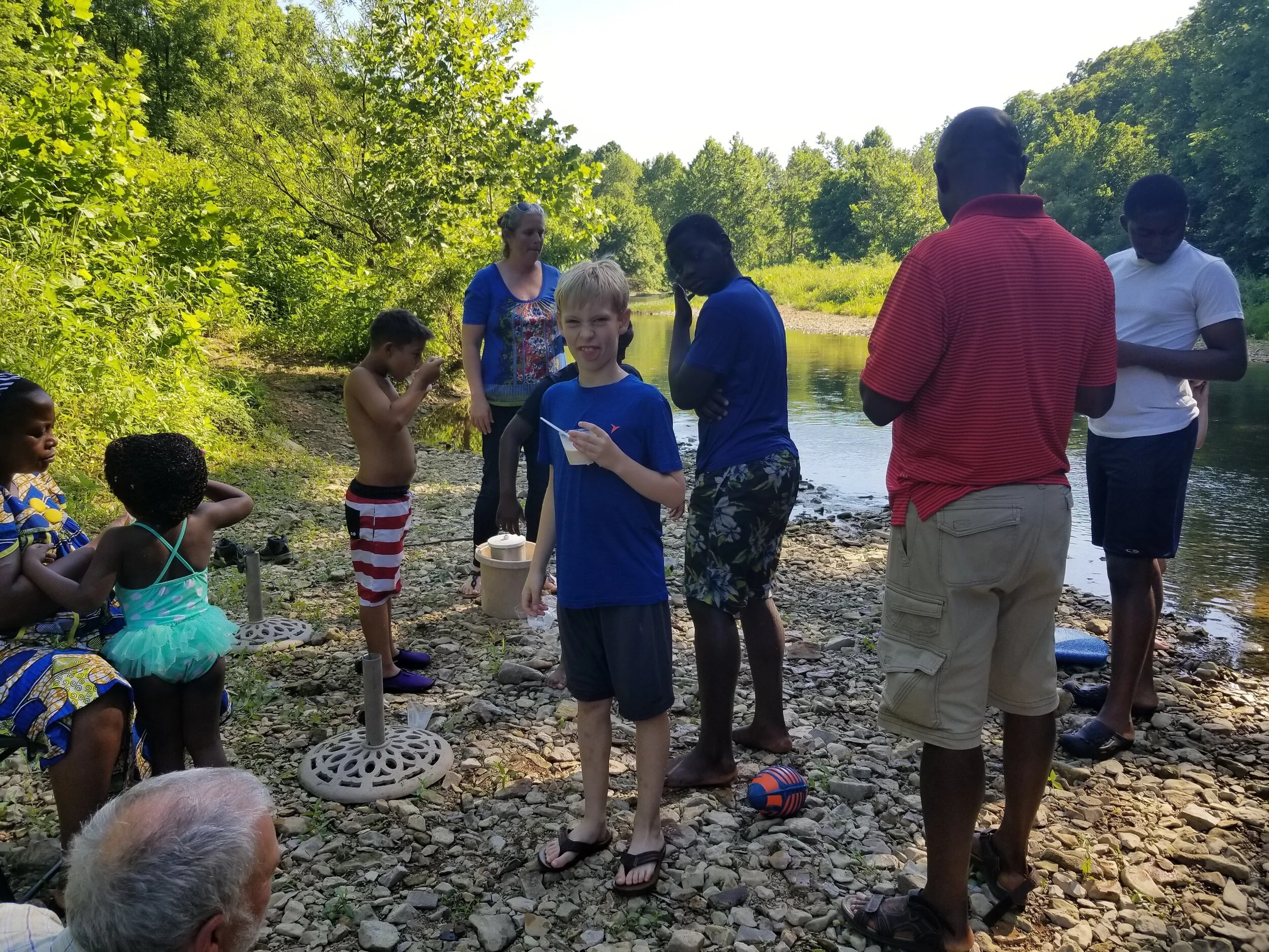 Group of people, including children and adults, standing on a rocky riverbank, surrounded by trees and greenery, in casual summer clothing. Some are interacting or eating.