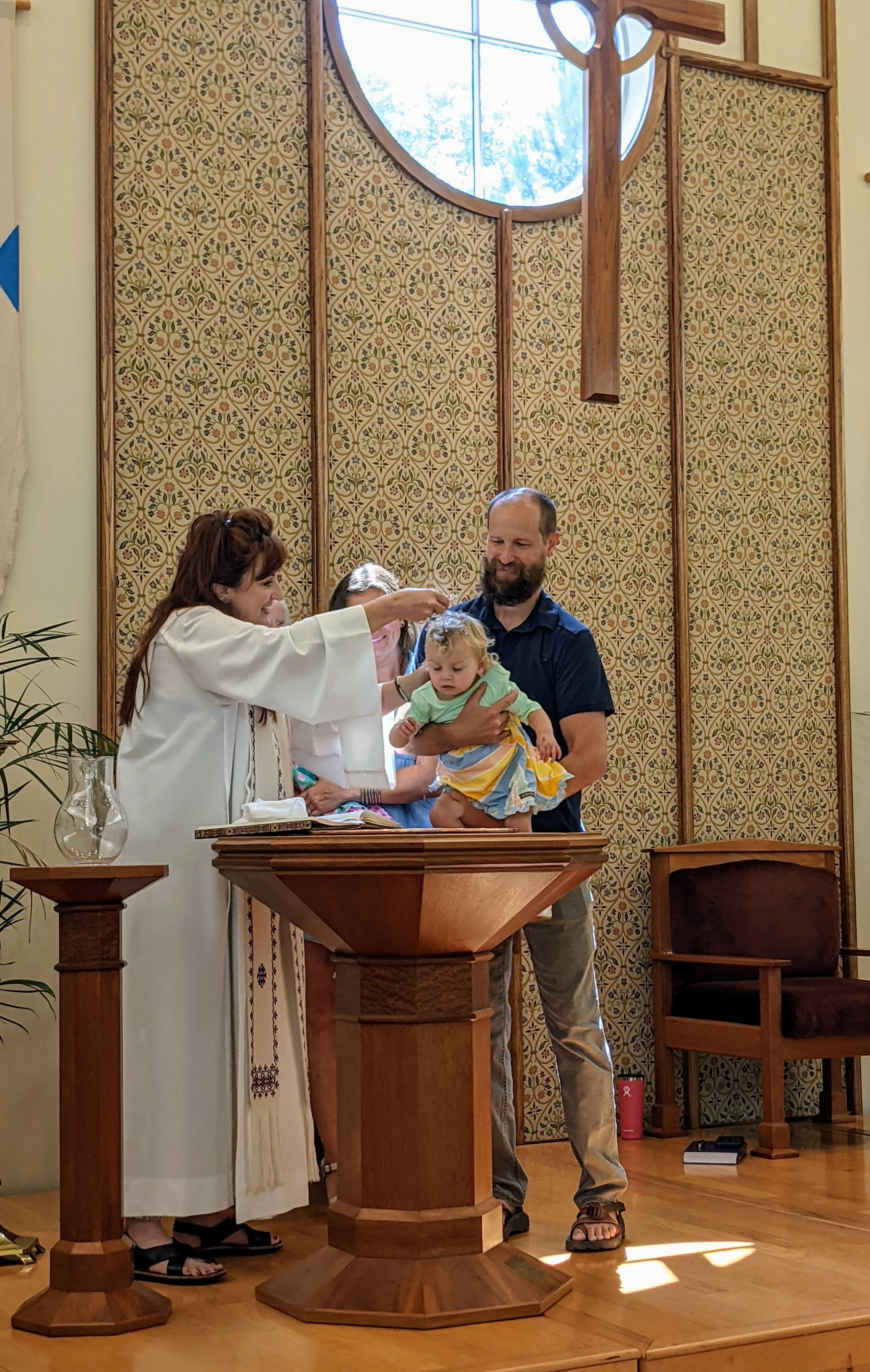 A religious baptism ceremony taking place in a church, featuring a priest in robes pouring water on a baby's head held by a man, with a woman standing nearby. The setting includes a baptismal font, decorative background, and a large wooden cross.