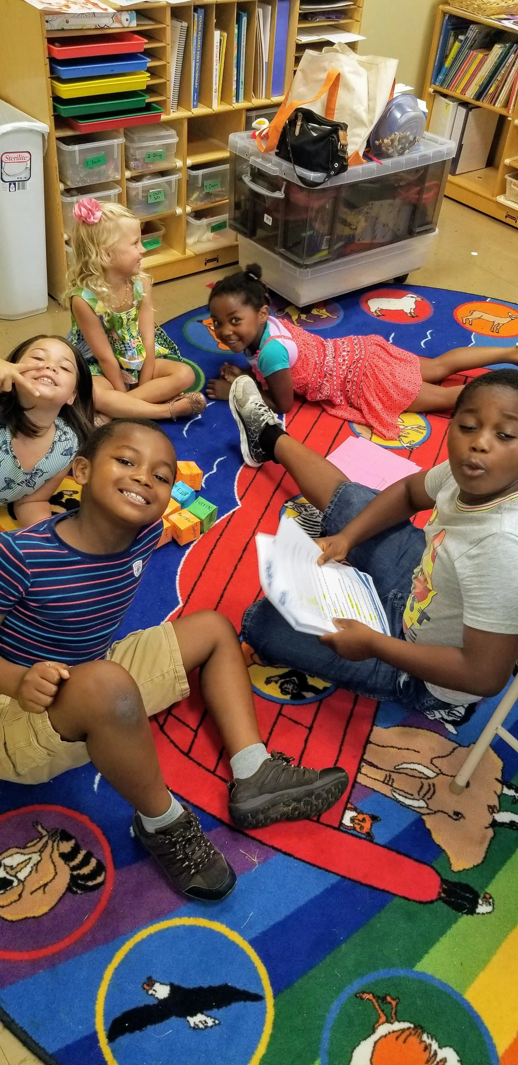 Children sitting on a colorful educational rug in a classroom, surrounded by shelves and storage containers, engaging in group activities.