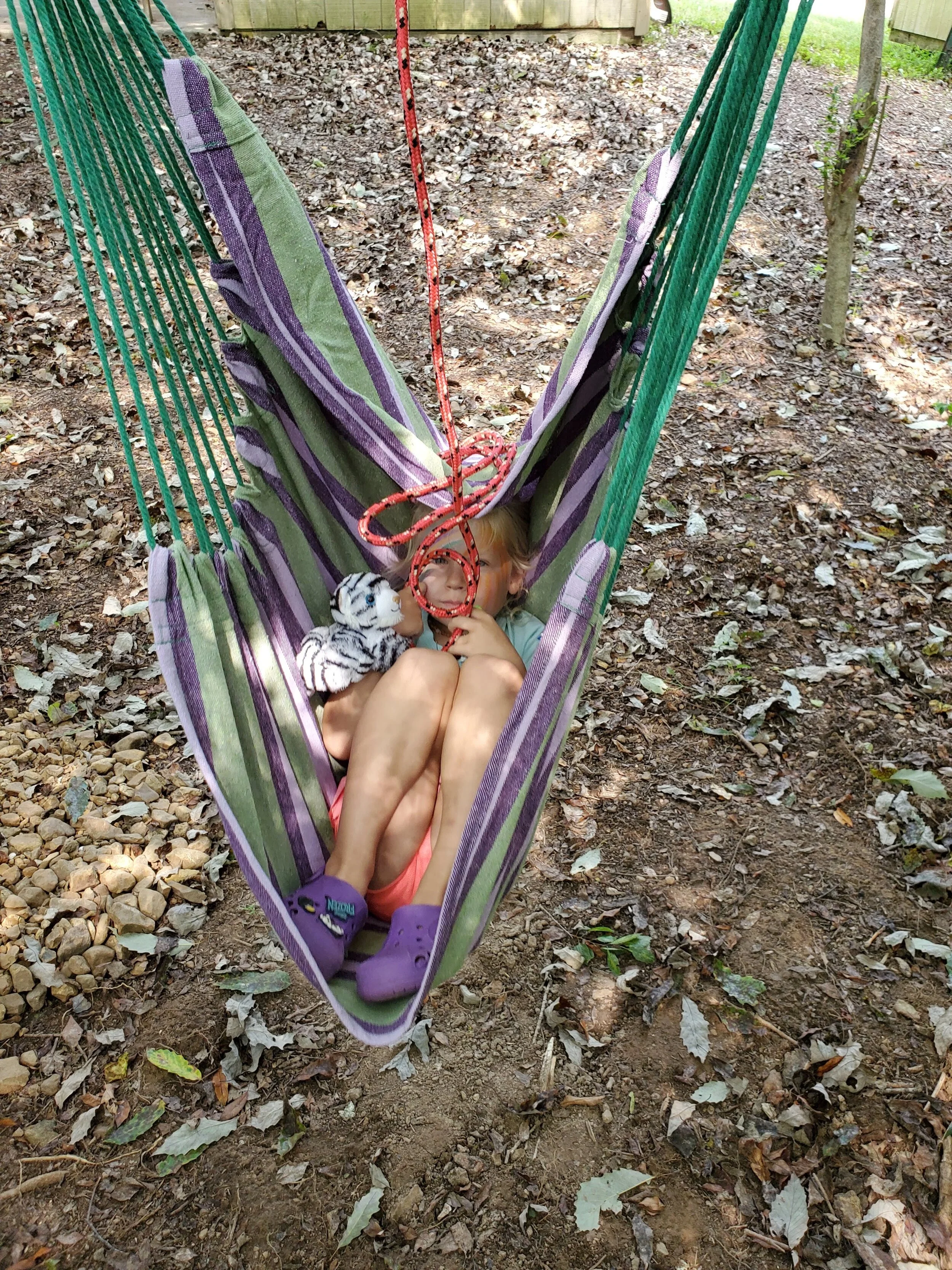 Child relaxing in a striped hammock with a stuffed toy outdoors.