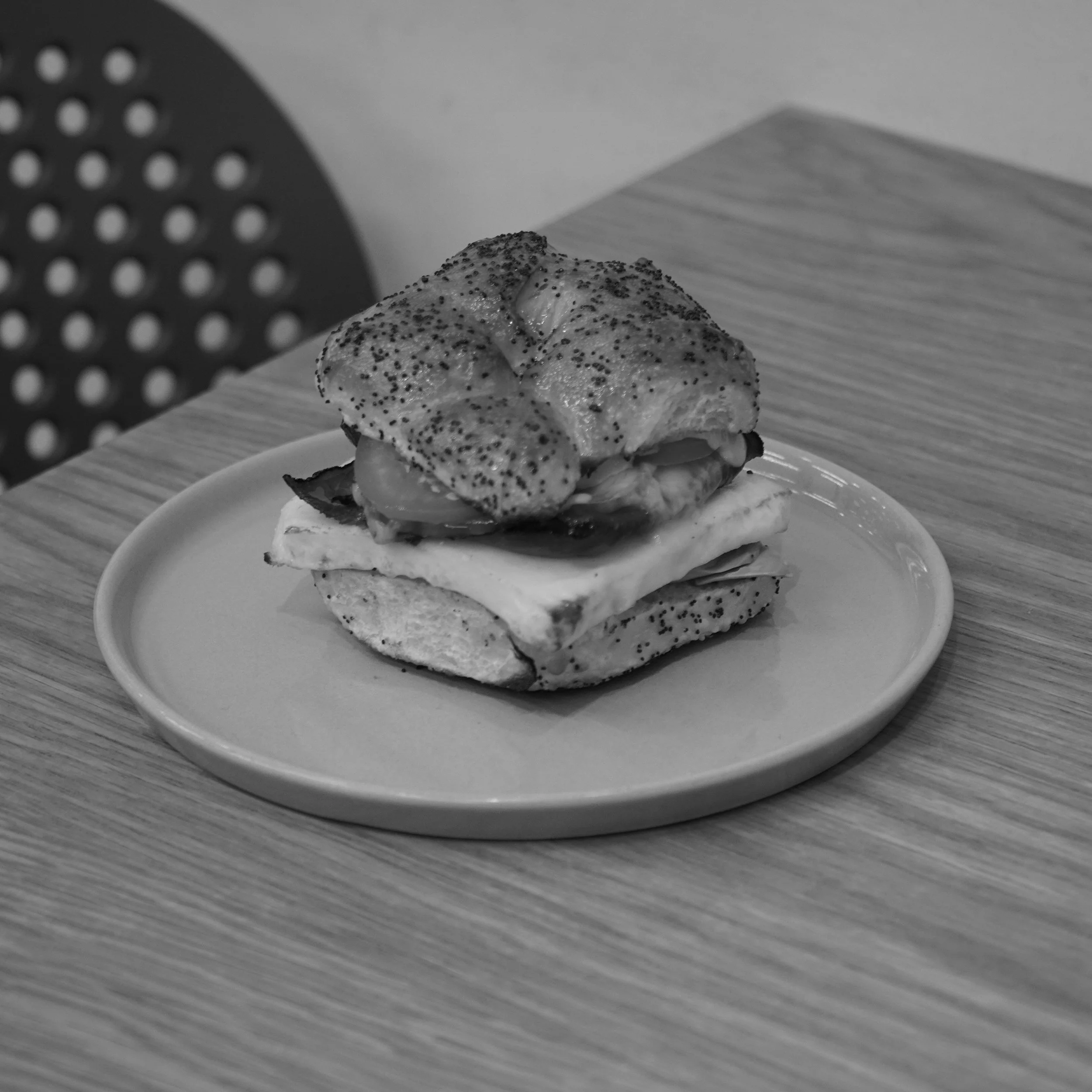 A sandwich with a poppy seed bun, lettuce, tomato, cheese, and a fried egg on a round plate on a wooden table.