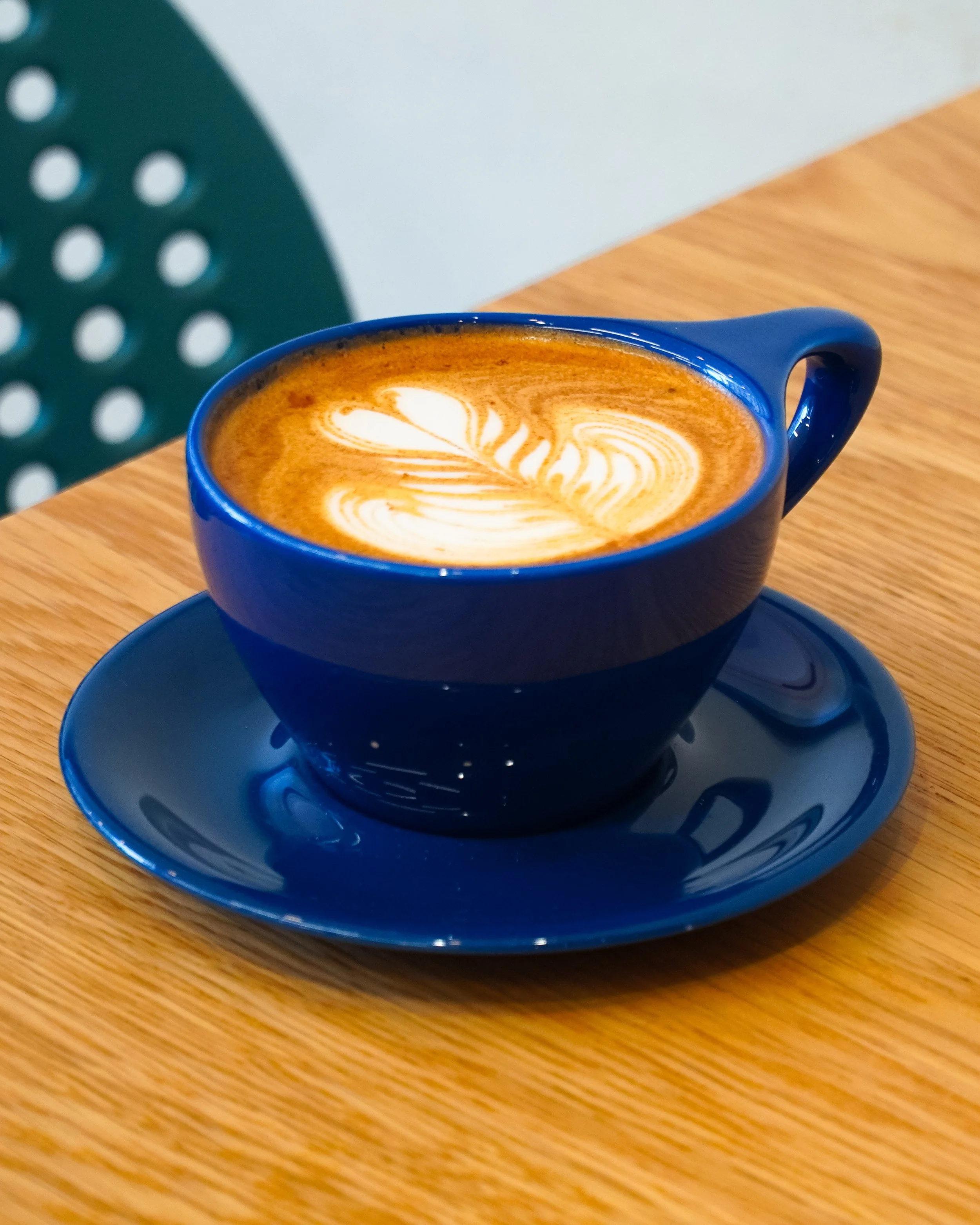 Blue ceramic cup of latte with latte art on top, placed on a matching blue saucer on a wooden table.