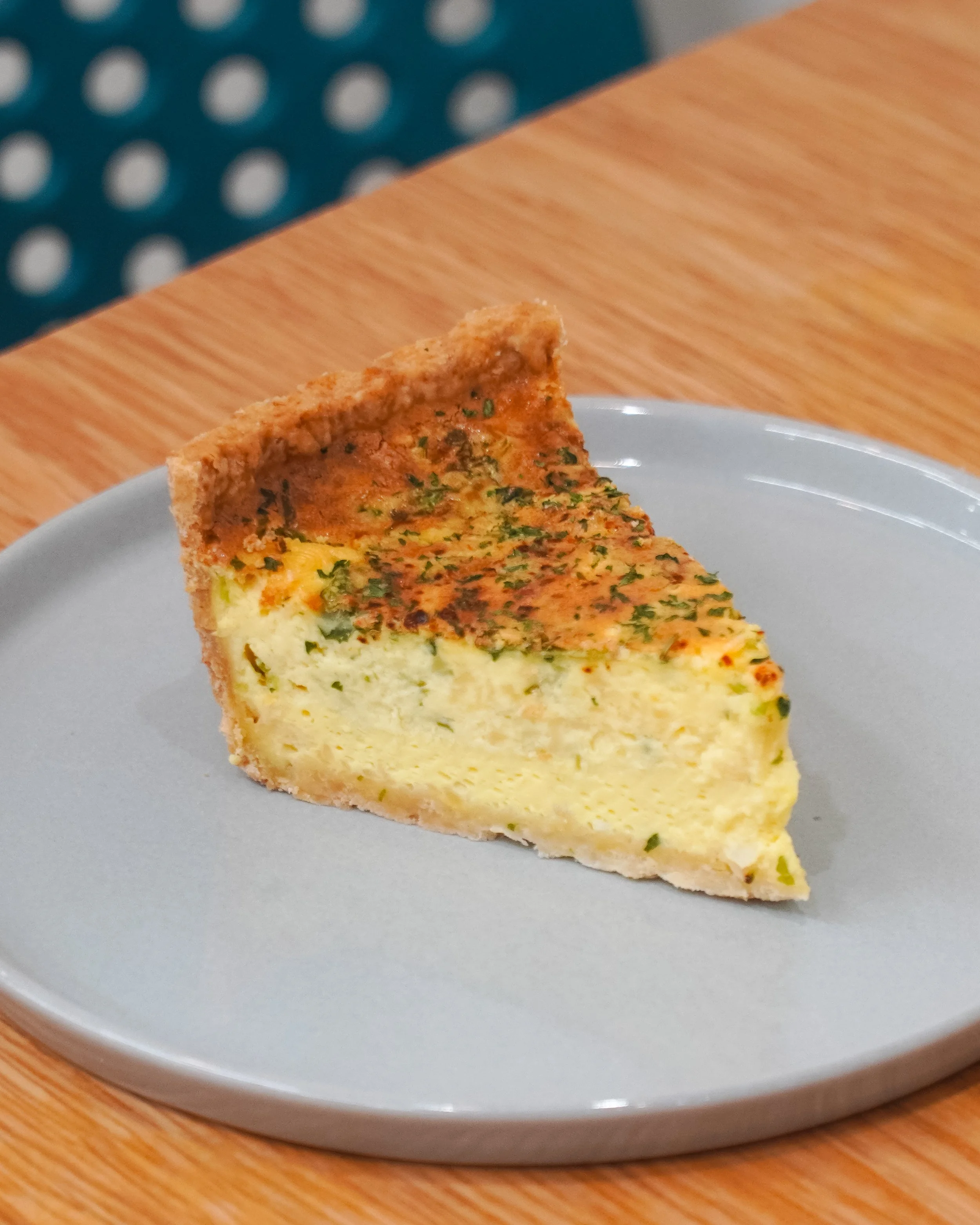 A slice of quiche on a gray plate, with a golden brown crust and creamy cheese filling, garnished with herbs.