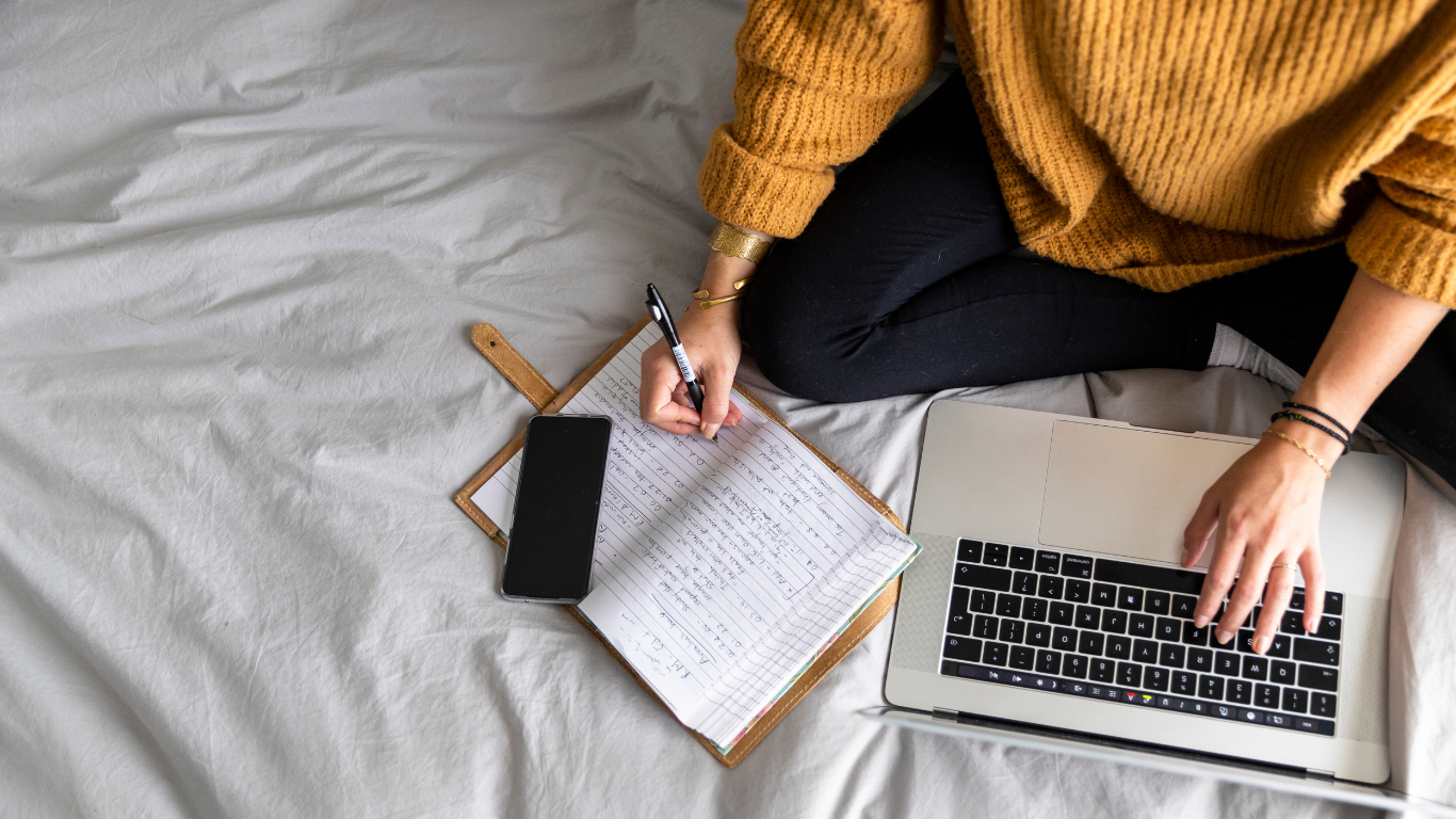 Person sat on bed and writing in journal with one hand writing and another hand on laptop keyboard
