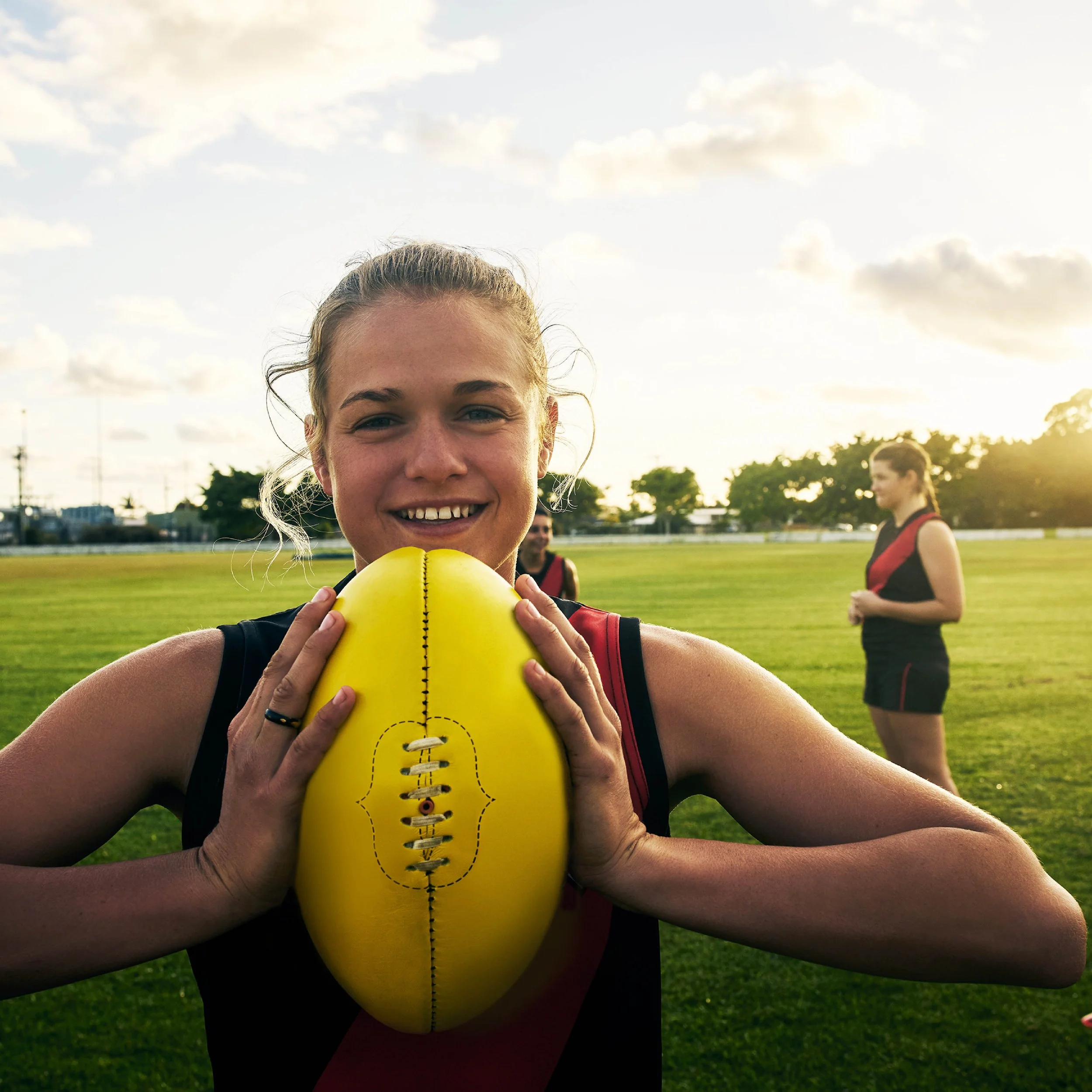 Close-up of a young woman in sports attire holding a yellow football, smiling, with other people in sport uniforms and a grassy field in the background during late afternoon or early evening.