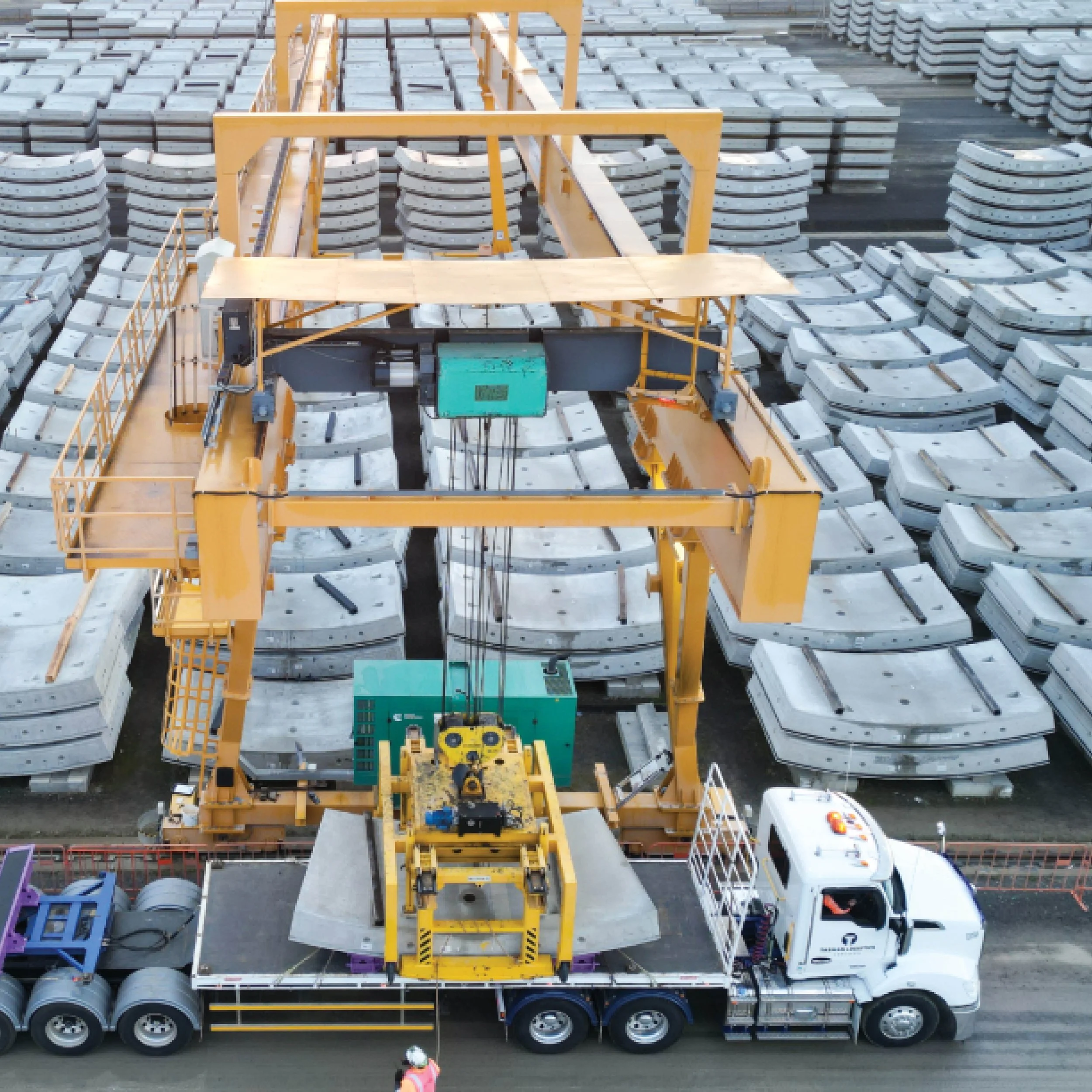 A construction crane lifting materials over a parking lot with trucks and equipment, surrounded by stacked concrete curbs.