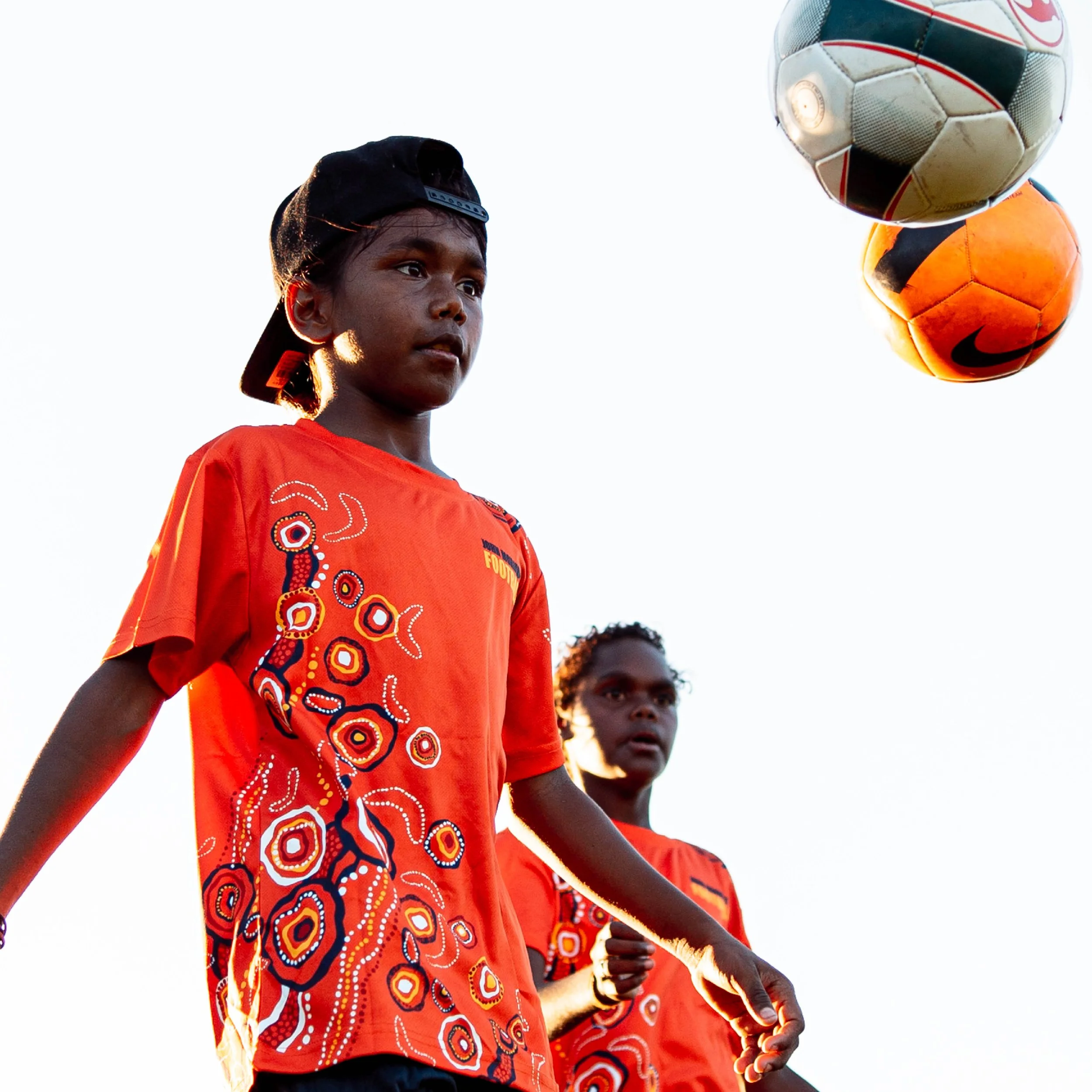 Young boy wearing a black cap and orange shirt with colorful pattern, playing soccer with two other children, against a white sky background.