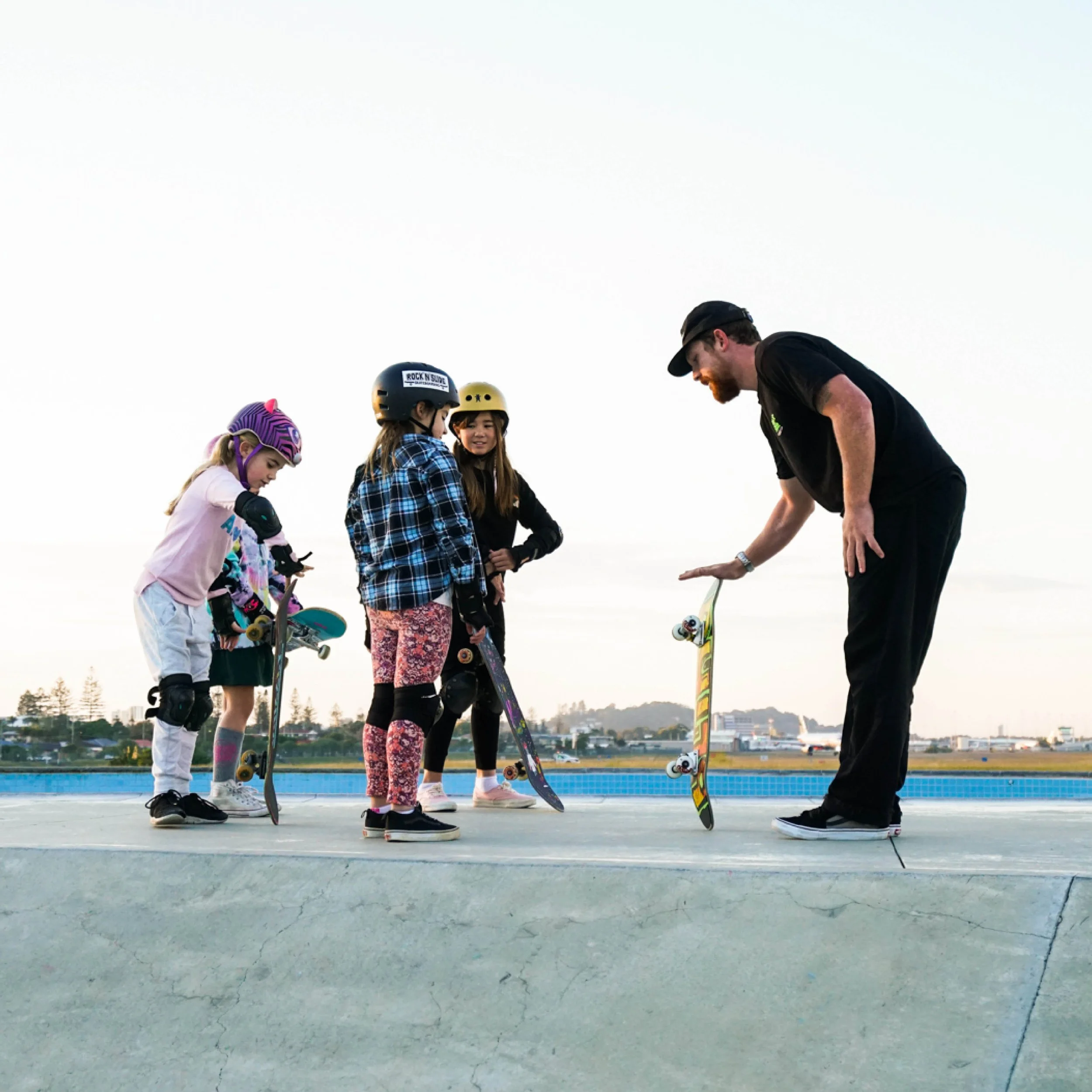 A group of children in skate helmets and knee pads standing on a concrete skate park ledge, listening to an adult instructor with a skateboard, under a clear sky with a cityscape in the background.