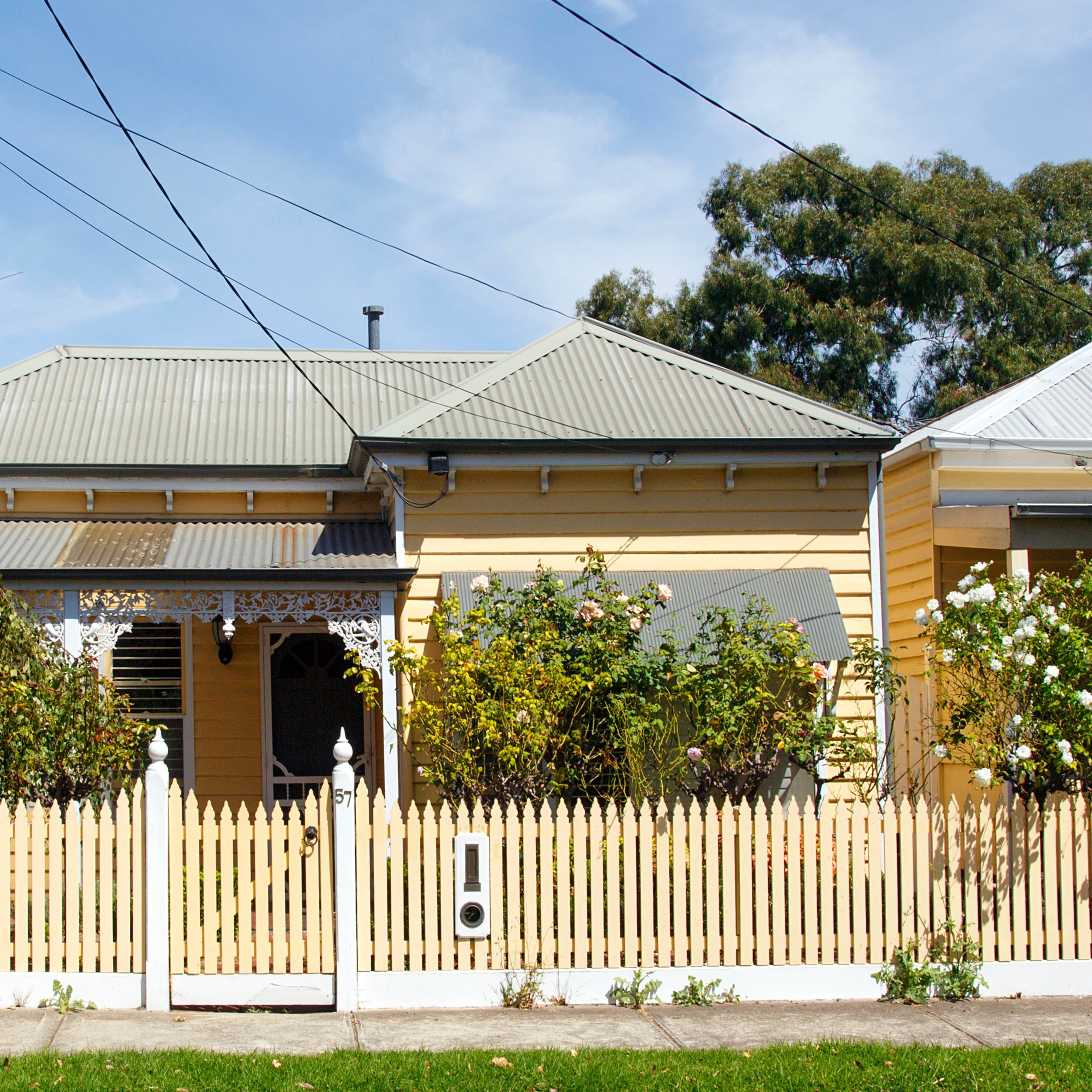 A house with a white picket fence, a chain link gate with a handle, a mailbox, a flower bed, and a yellow siding exterior. There are trees and a blue sky with some clouds in the background.