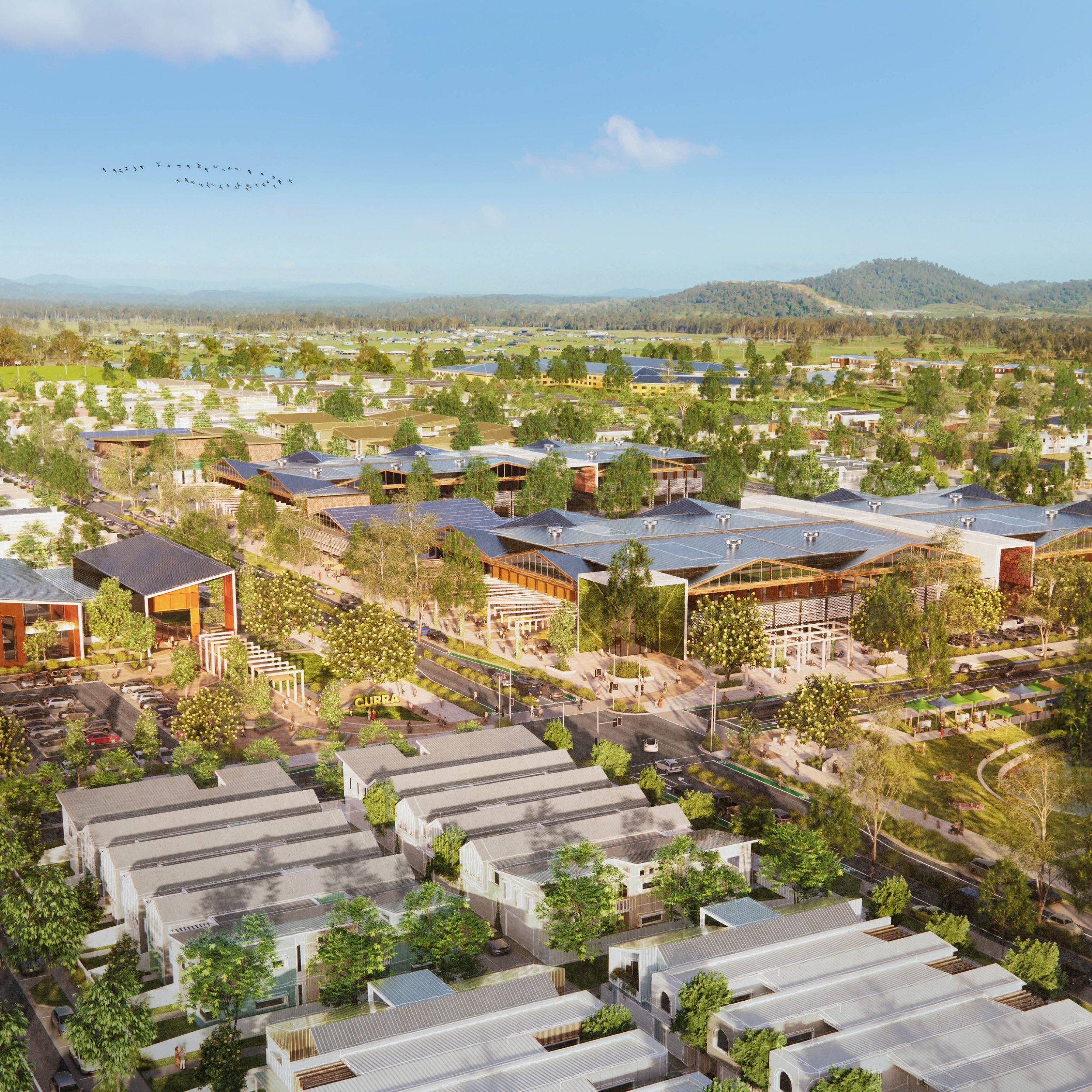 Aerial view of a suburban area with residential houses, trees, parking lots, and a park with a 'CURA' sign, set against a backdrop of green hills and a blue sky with some clouds.