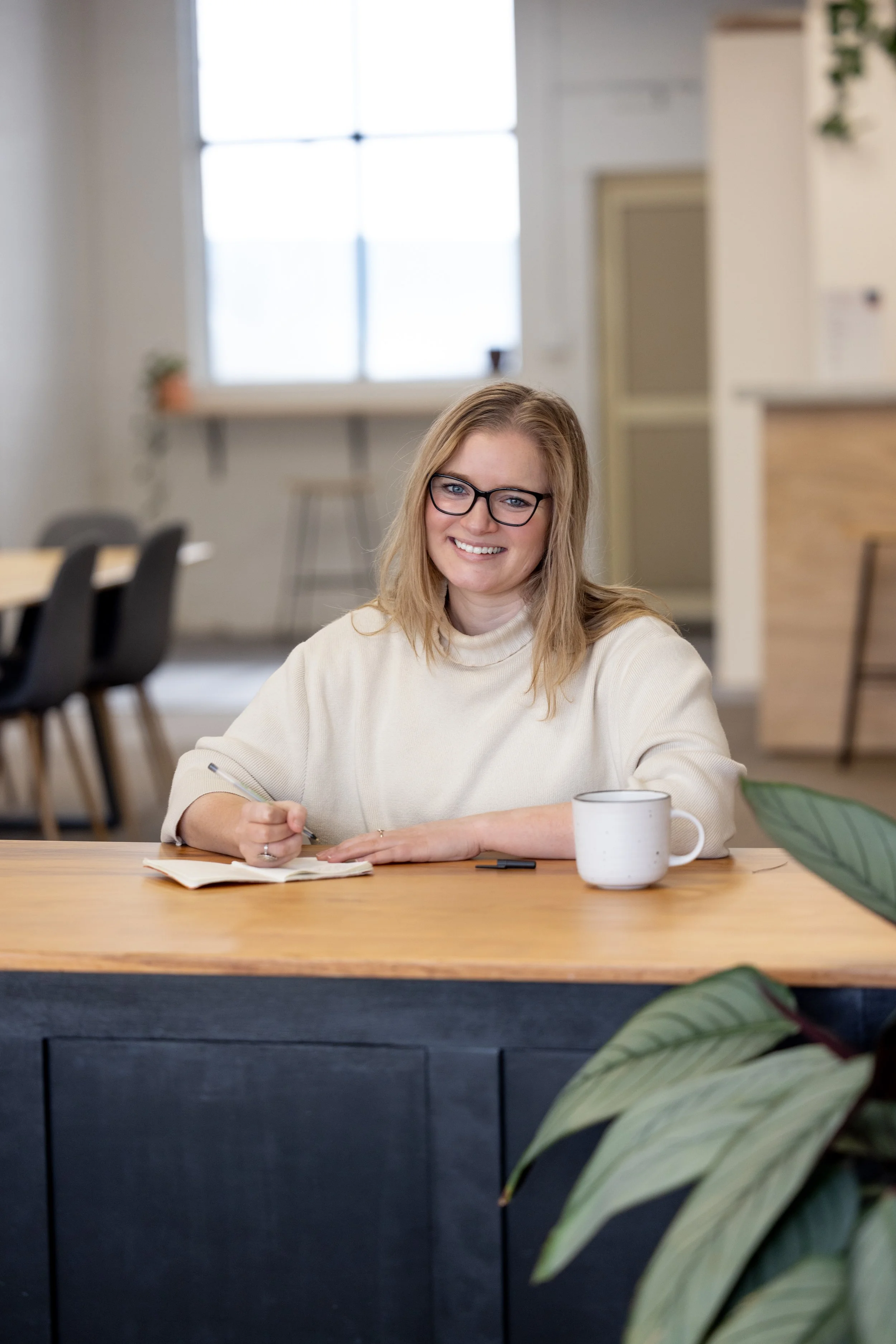 A woman with blonde hair, glasses, and a cream sweater smiling while sitting at a wooden table, writing in a notebook with a white mug nearby in a bright, modern office or café setting.