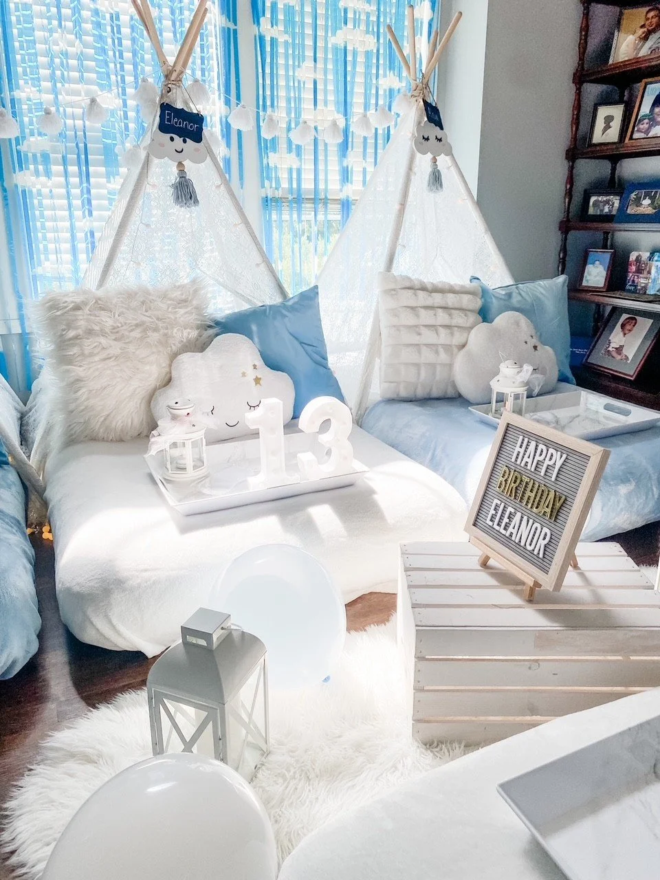 Decorated rooms with white and blue tones for a child's birthday celebration, featuring teepees, fluffy pillows, cloud-shaped cushions, balloons, a lantern, and a birthday sign reading 'Happy Birthday Eleanor'.